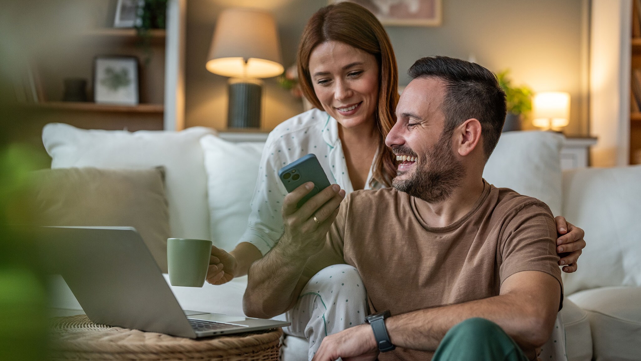 Eine Frau und ein Mann sitzen vor einem Laptop. Der Mann hält ein Smartphone in der Hand.