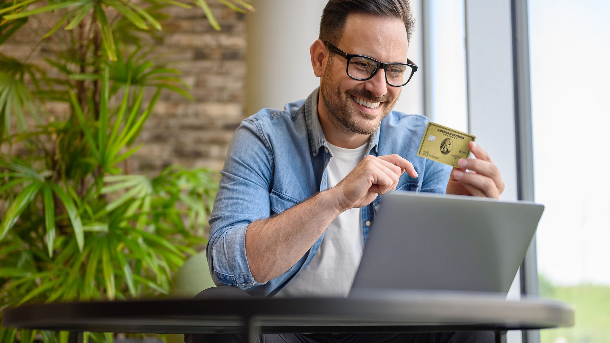 Ein Mann sitzt vor seinem Laptop und hält eine goldene Kreditkarte von American Express in der Hand.