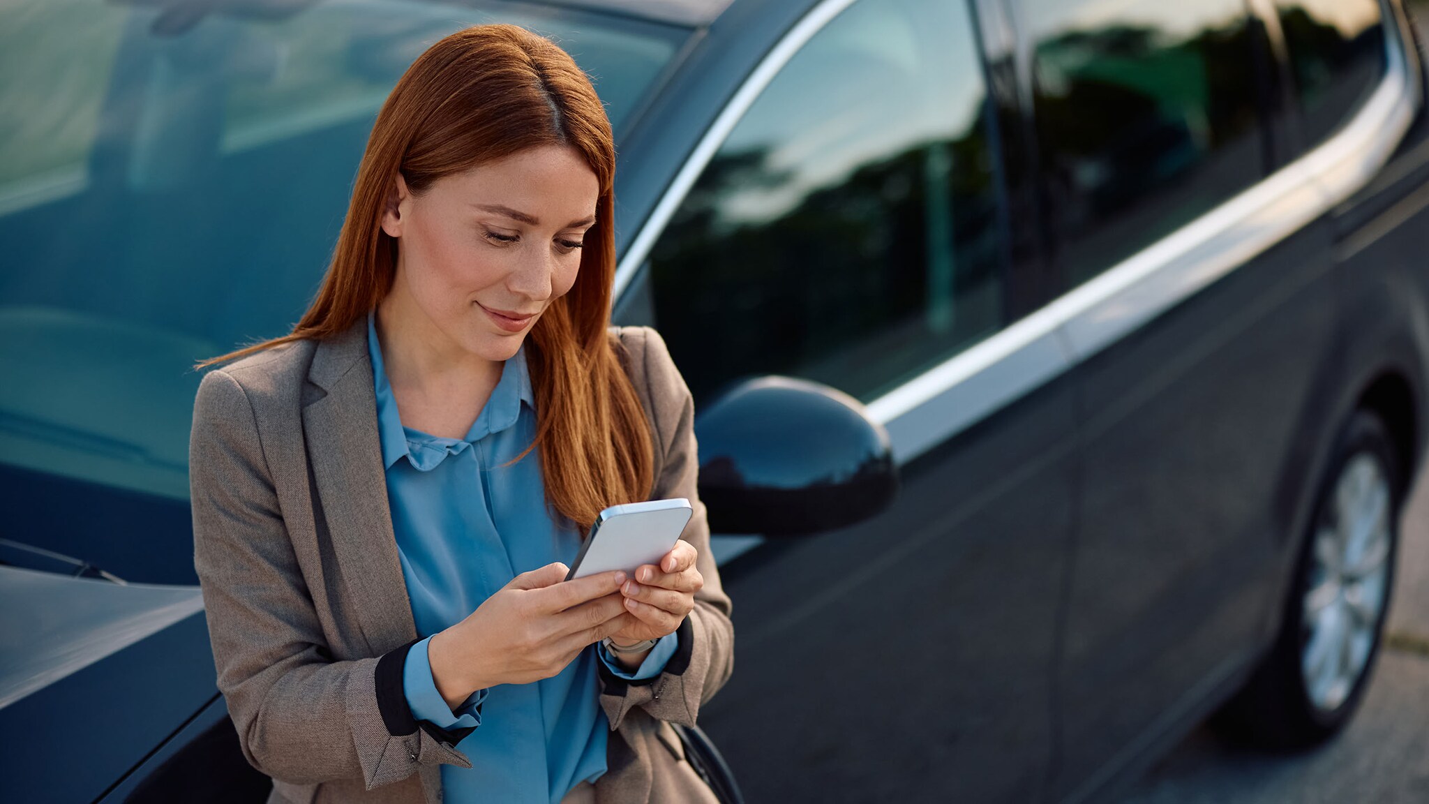 Eine Frau lehnt an einem Auto und schaut auf ihr Smartphone.