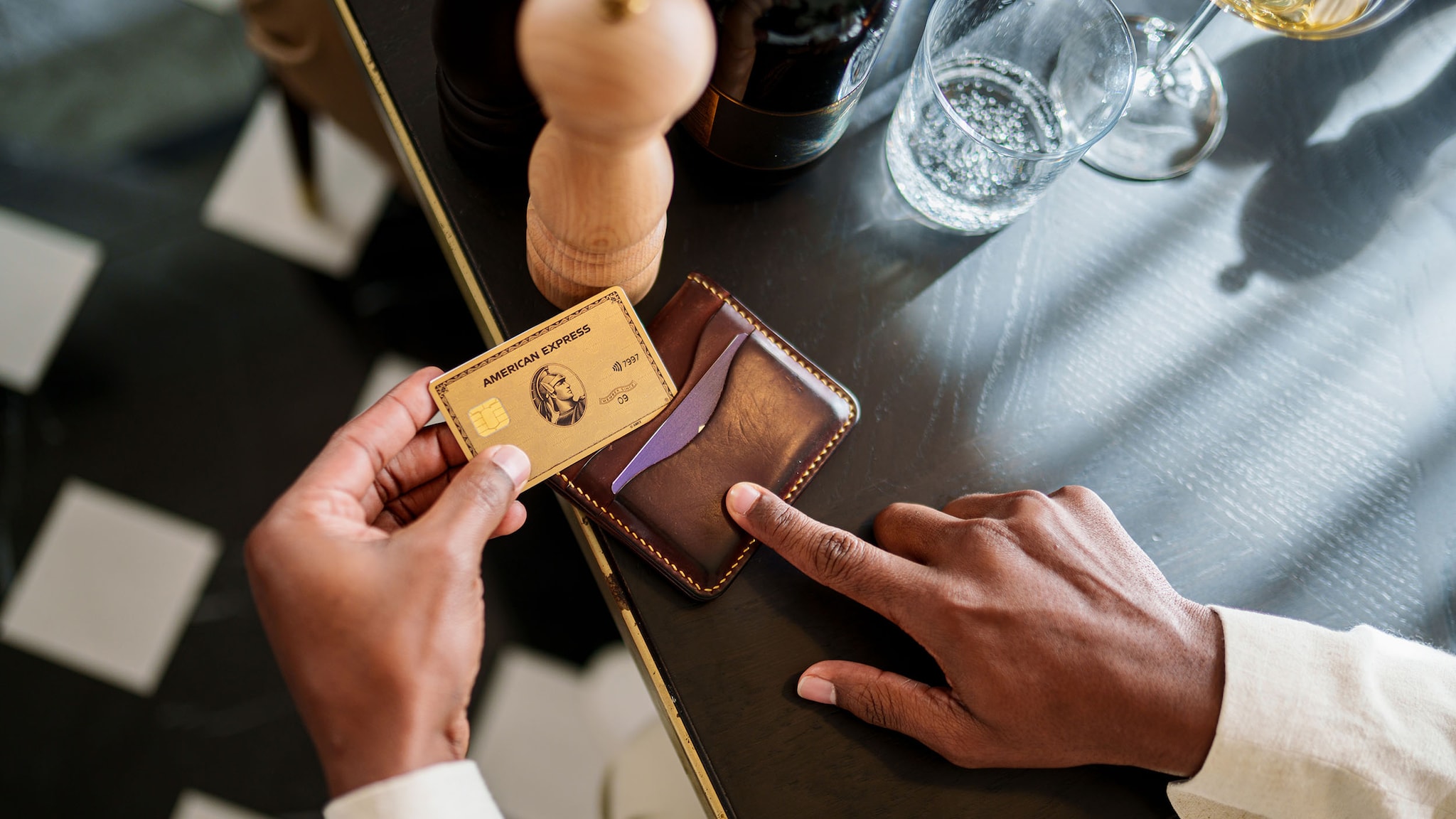 Aufsicht einer goldenen Kreditkarte von American Express, die von einer Hand aus einem Lederetui gezogen wird auf einem Restauranttisch.