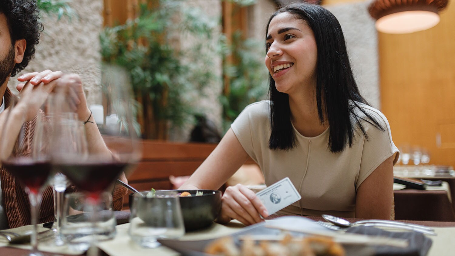 Eine Frau sitzt gut gelaunt an einem Tisch im Restaurant mit weiteren Personen und hält eine silberne Kreditkarte von American Express in der Hand.