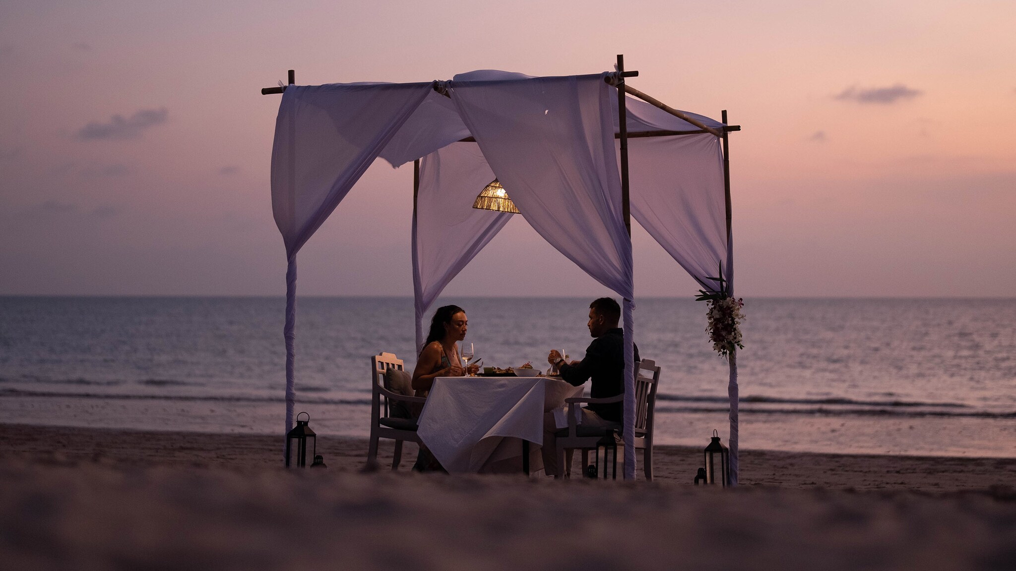 Ein Mann und eine Frau sitzen beim Dinner an einem Tisch unter einem Baldachin am Strand im Abendrot.