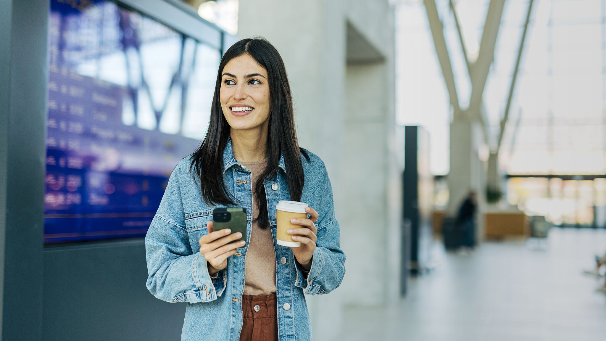 Eine Frau steht mit Kaffeebecher und Smartphone in den Händen in einem Flughafenterminal.