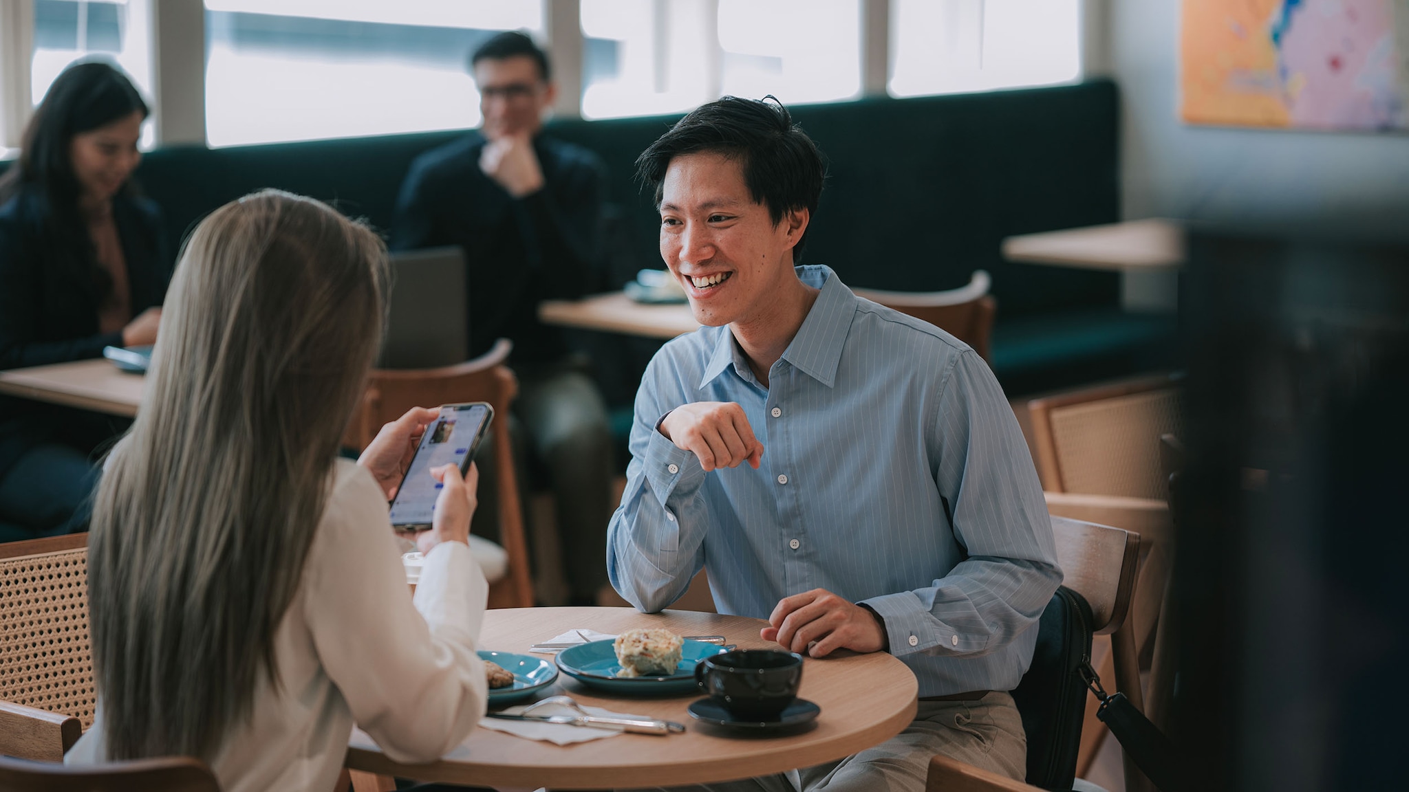 Ein Mann und eine Frau sitzen sich gegenüber an eienm Bistrotisch in einer Lounge mit weiteren Personen, sie hält ein Smartphone in ihren Händen.