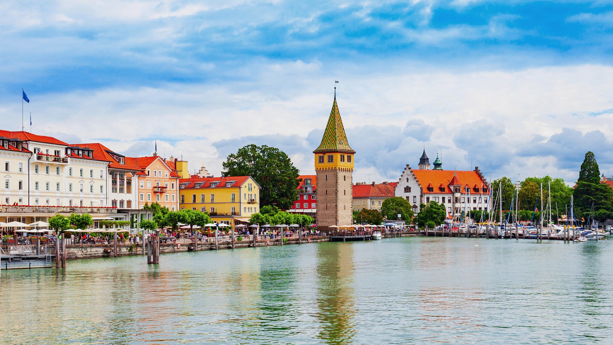 Hafenpromenade mit historischen Häusern und mittelalterlichem Turm bei Sonnenschein, im Vordergrund Wasser.