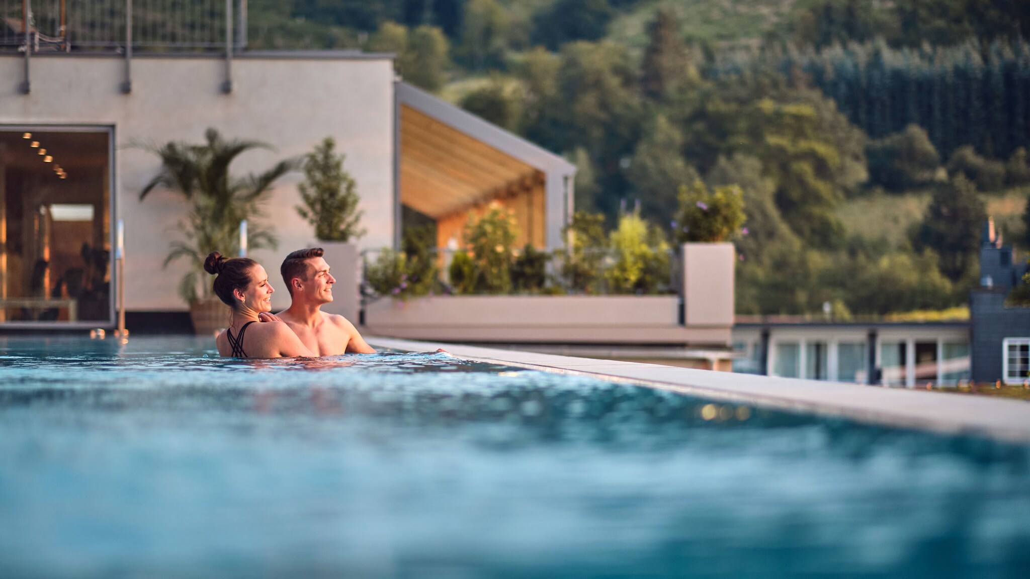 : Eine Frau und ein Mann stehen beieinander in einem Infinity-Pool an einem Gebäude und blicken in die Ferne vor einem bewaldeten Berghang.
