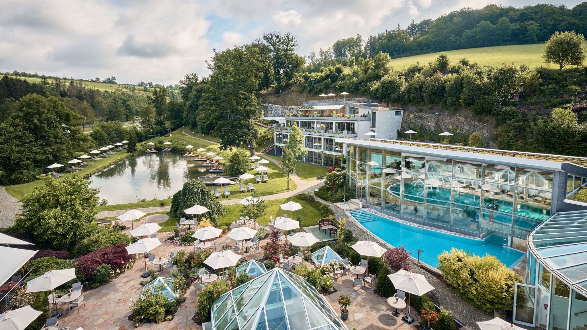Garten einer luxuriösen Hotelanlage mit Sonnenschirmen an Pool und Teich, umgeben von hügeliger Waldlandschaft.