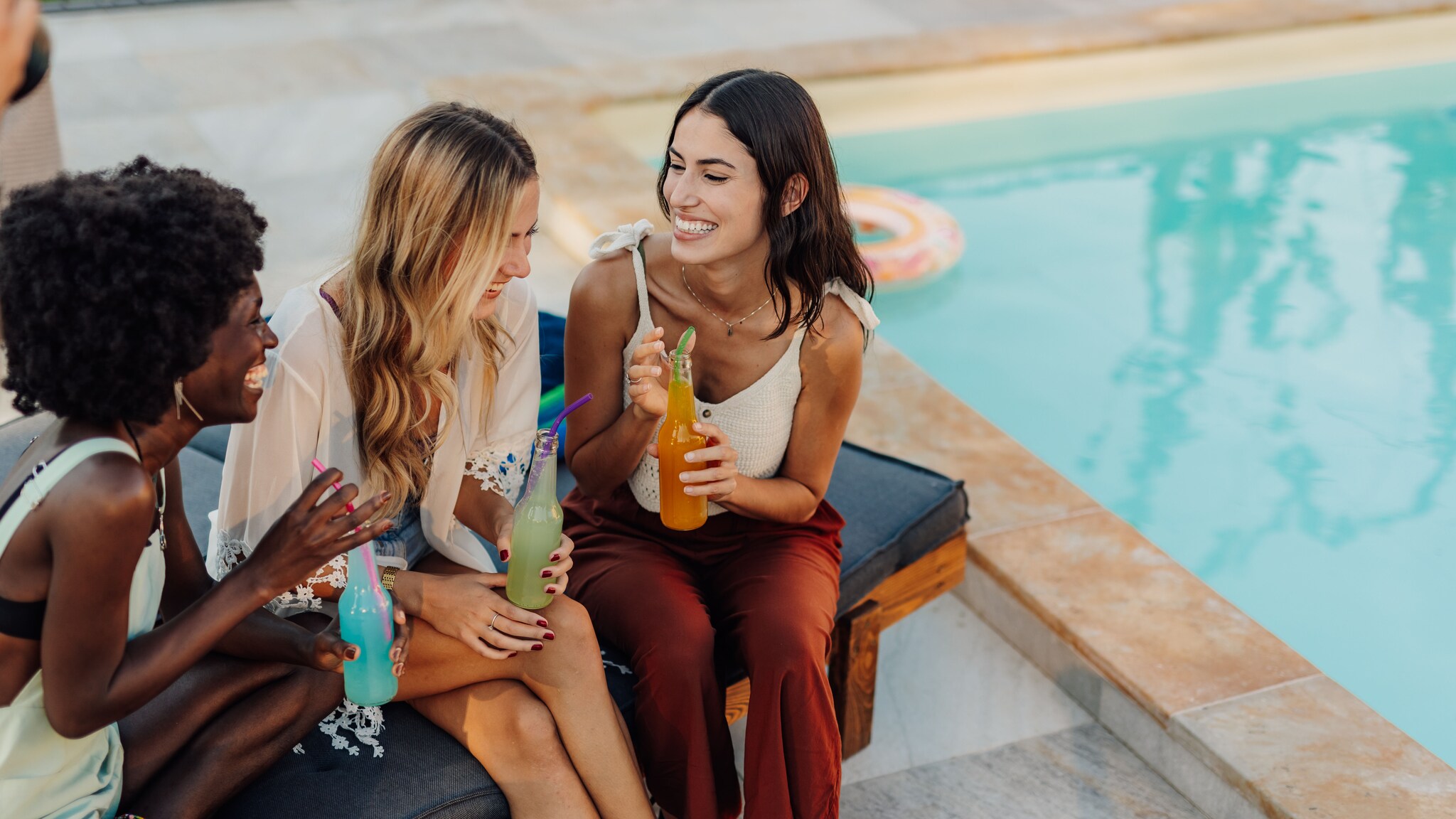 Drei junge Frauen sitzen mit einem Getränk in der Hand an einem Hotelpool.