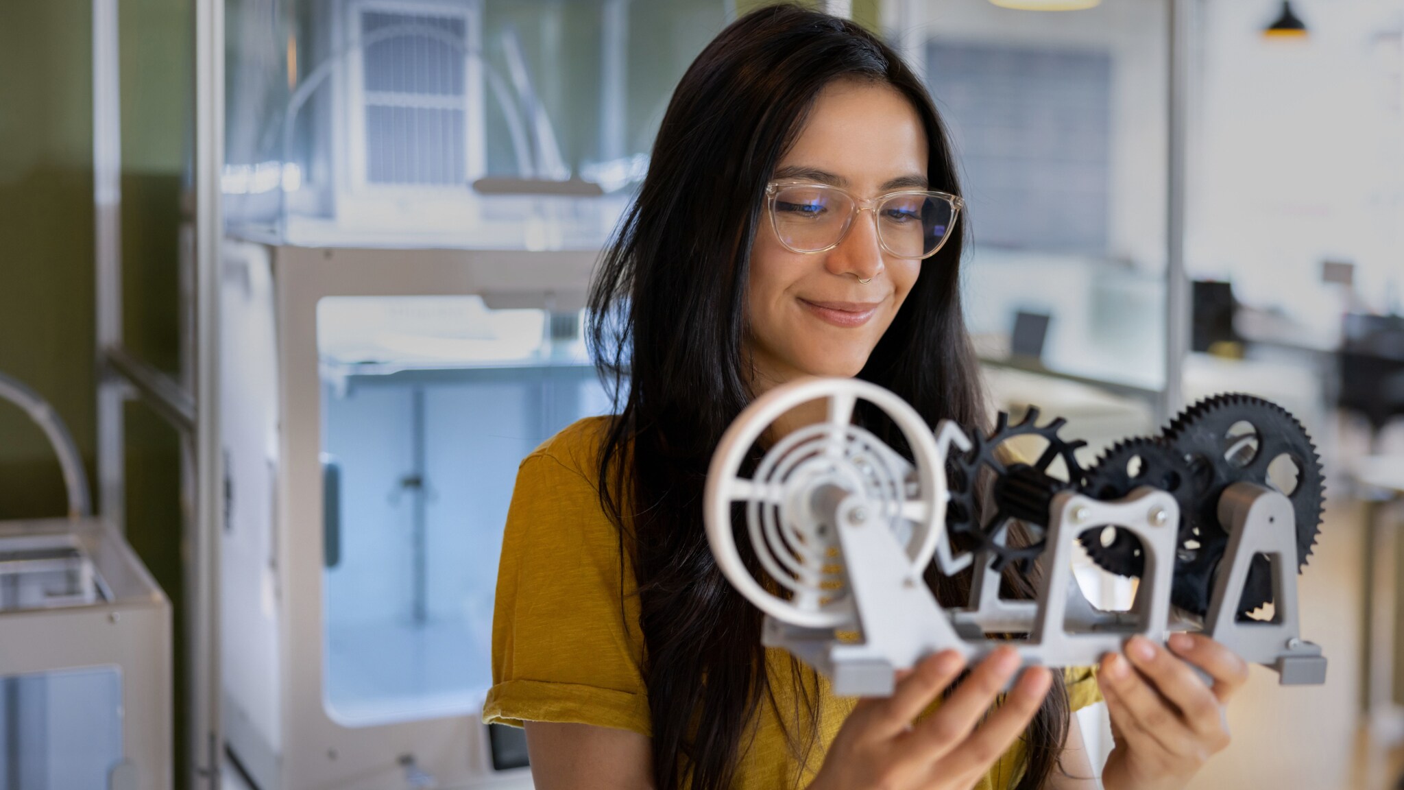 Eine Frau hält zufrieden lächelnd ein Modell mit Radgetrieben aus einem 3D-Drucker vor sich hoch in einem Labor.