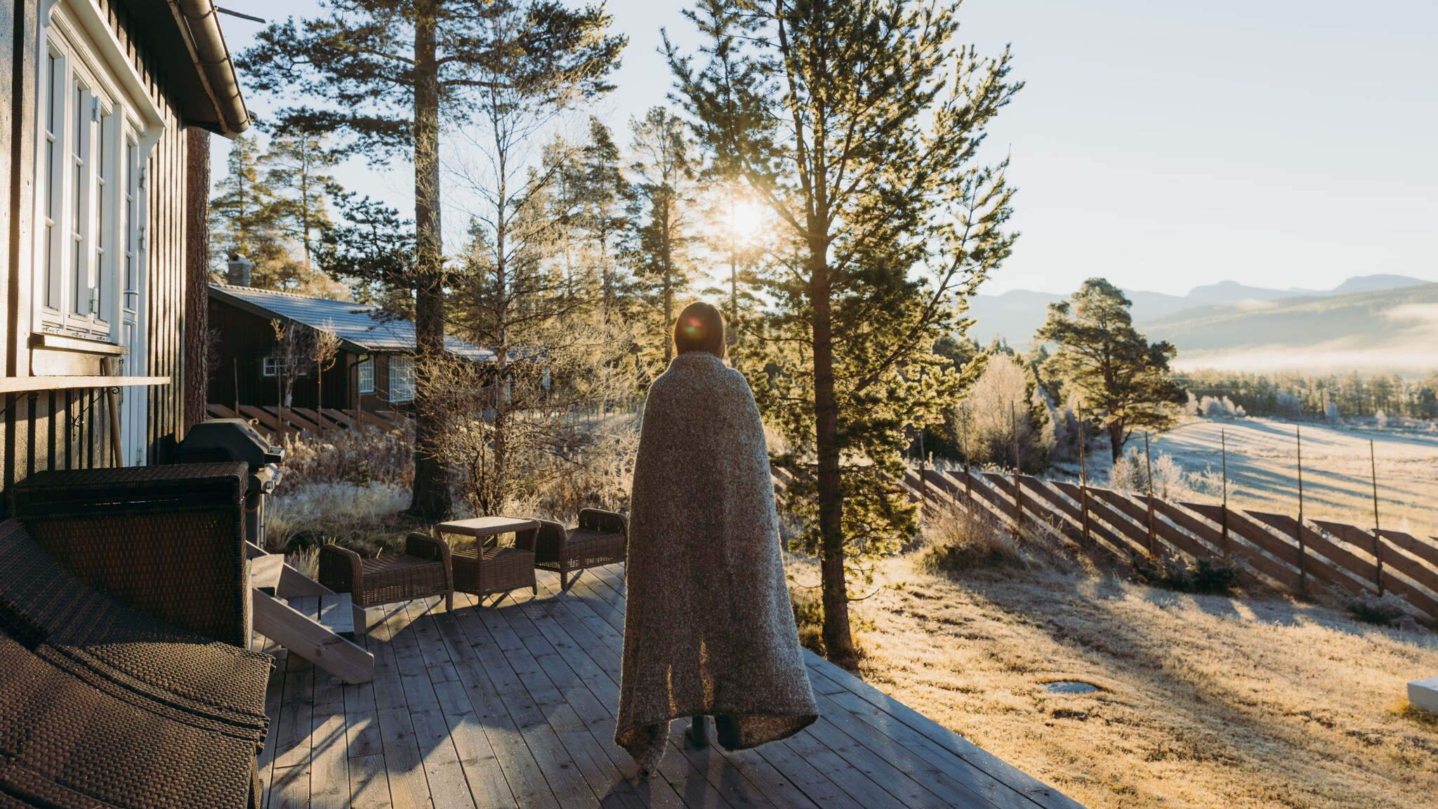 Rückansicht einer Person, die in eine Decke gehüllt auf der Holzterrasse vor einem Haus steht, im Hintergrund neblige Landschaft und Bäume.