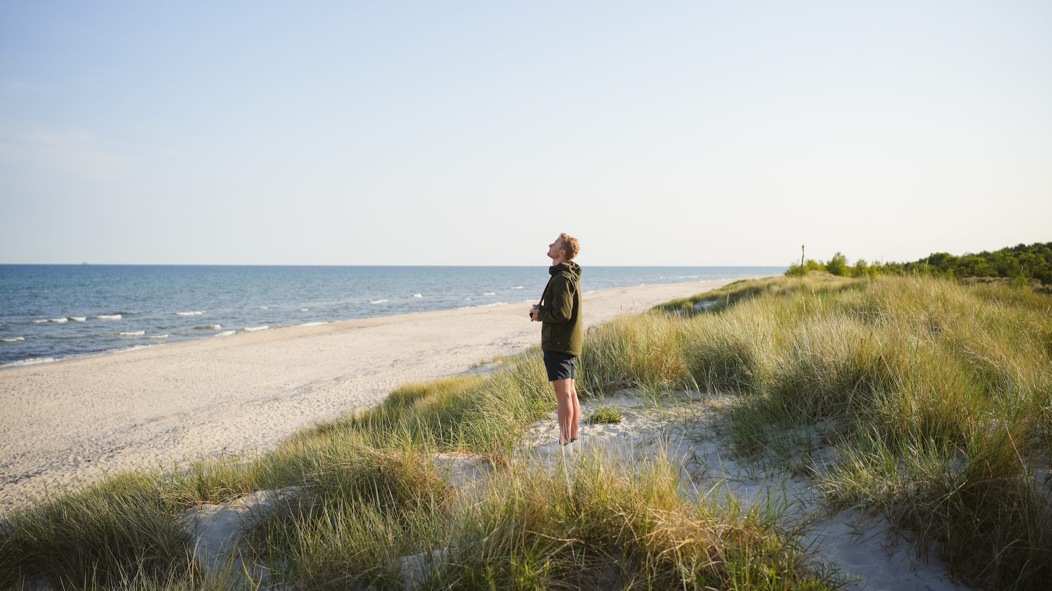 Ein Mann steht an einem weiten Sandstrand zwischen Gräsern und hat seinen Blick auf das Meer gerichtet.