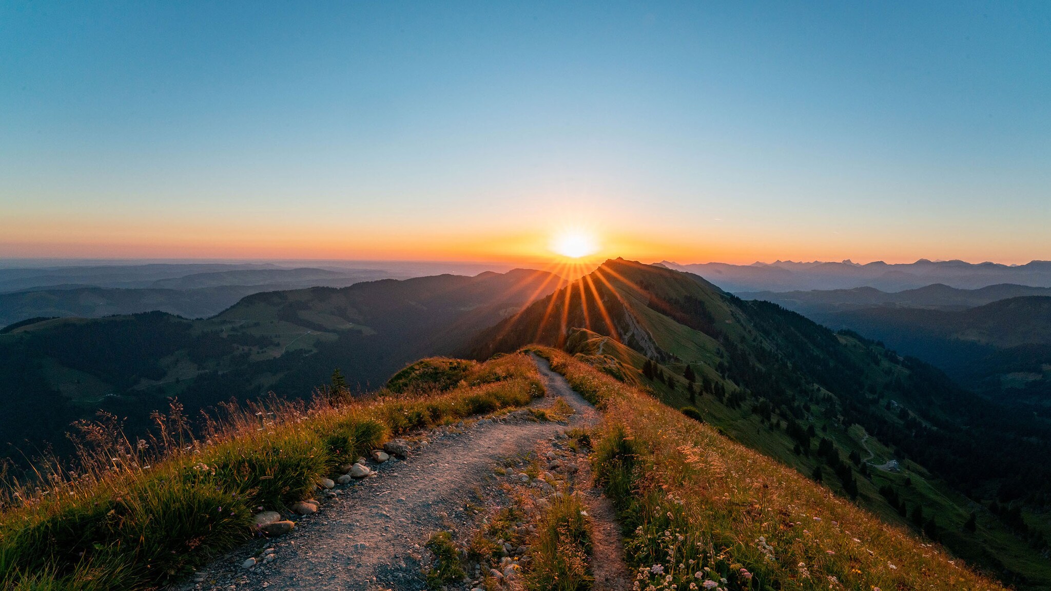 Auf einem Berggipfel liegt ein kleiner Wanderweg, im Hintergrund geht die Sonne unter.
