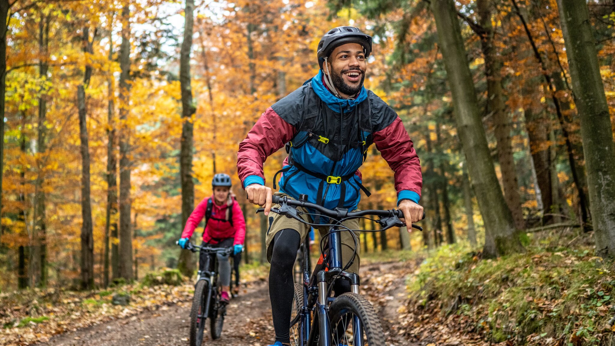 Zwei Personen in Sportbekleidung und mit Helmen, die auf Fahrrädern durch einen herbstlichen Wald fahren.
