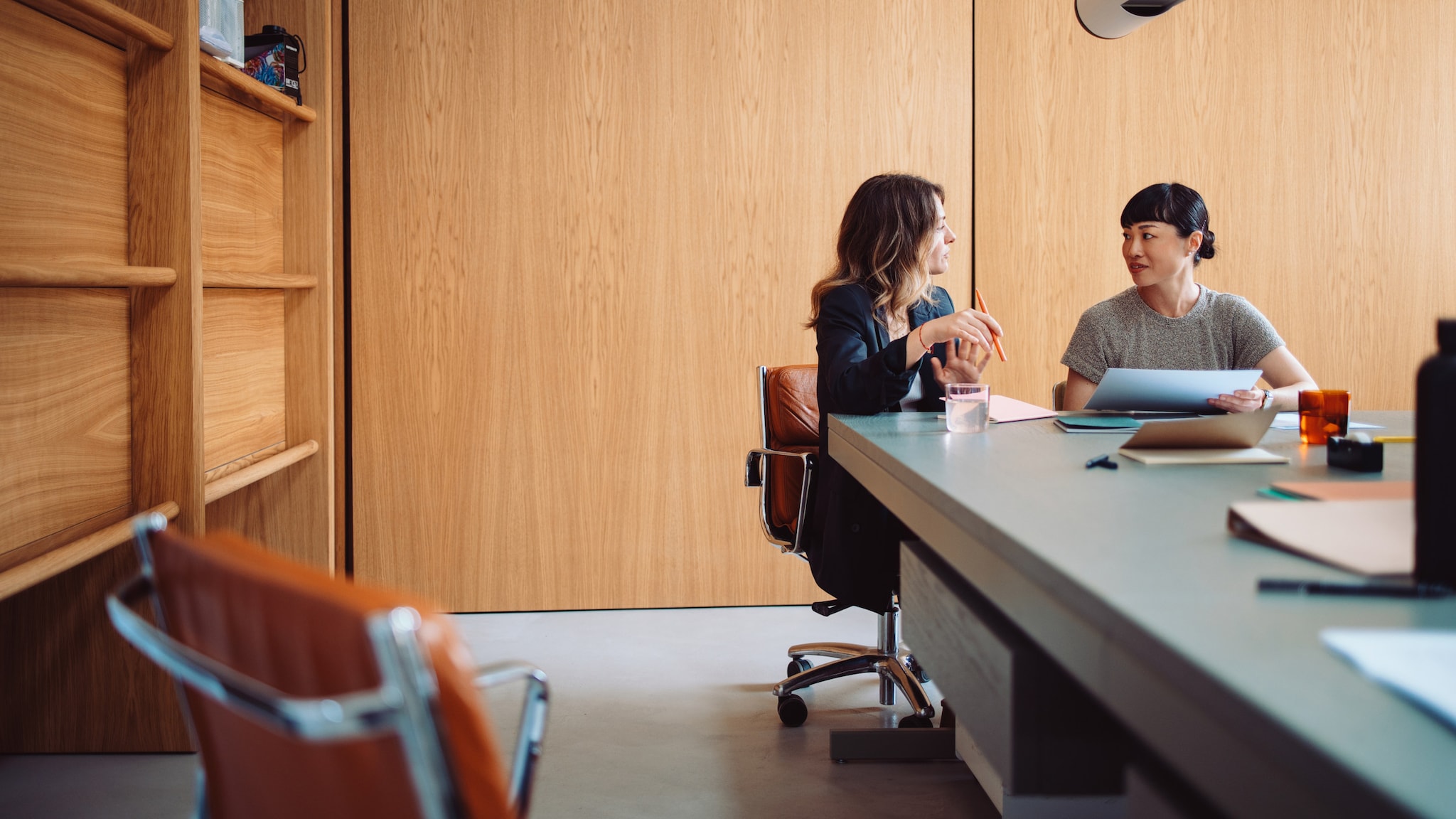 Zwei Geschäftsfrauen sitzen in einem Büro an einem langen Schreibtisch und unterhalten sich.
