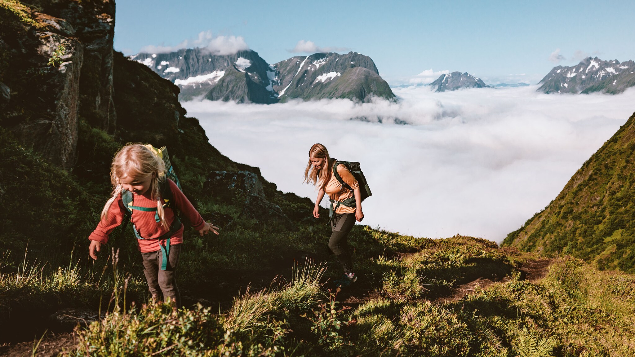 Ein kleines Mädchen und eine Frau laufen mit Rucksäcken einen Berg hinauf vor wolkenverhangenen Gipfelpanorama bei Sonnenschein.