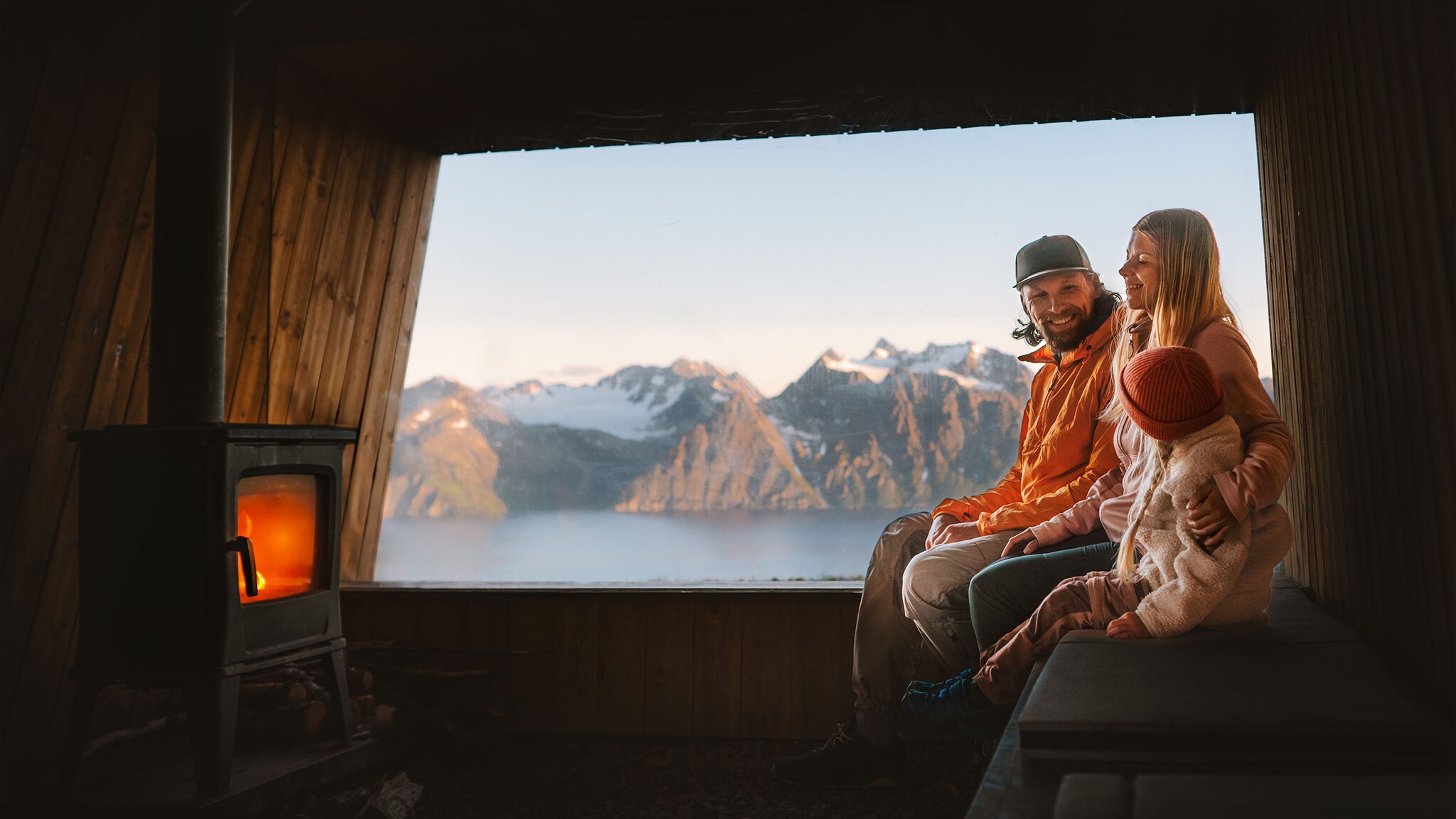 Ein Mann, eine Frau und ein Kind sitzen nebeneinander in einer Holzhütte mit Kaminfeuer vor einem Panoramafenster mit Blick auf einen See vor schneebedecktem Bergpanorama. 