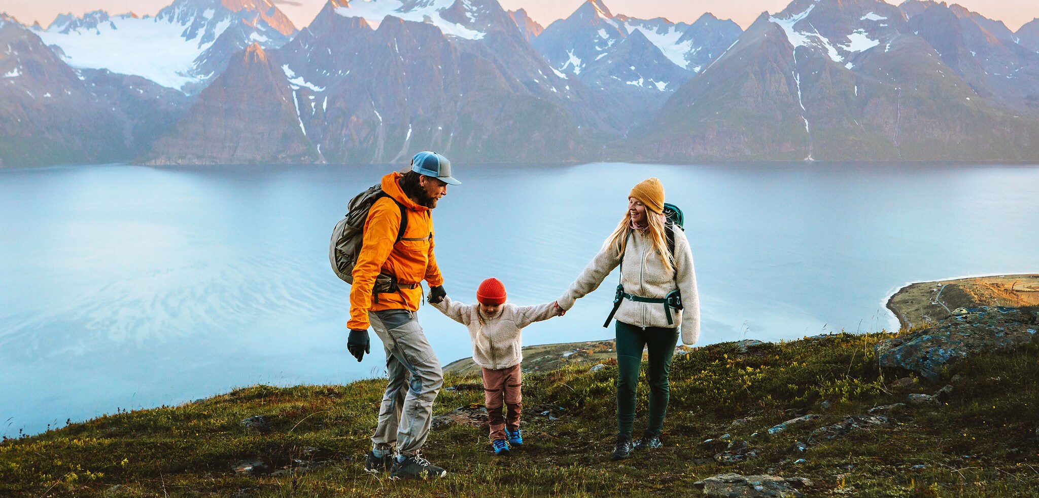 Ein Mann und eine Frau mit Rucksäcken halten ein Kind zwischen ihnen an den Händen an einem See vor Bergpanorama.