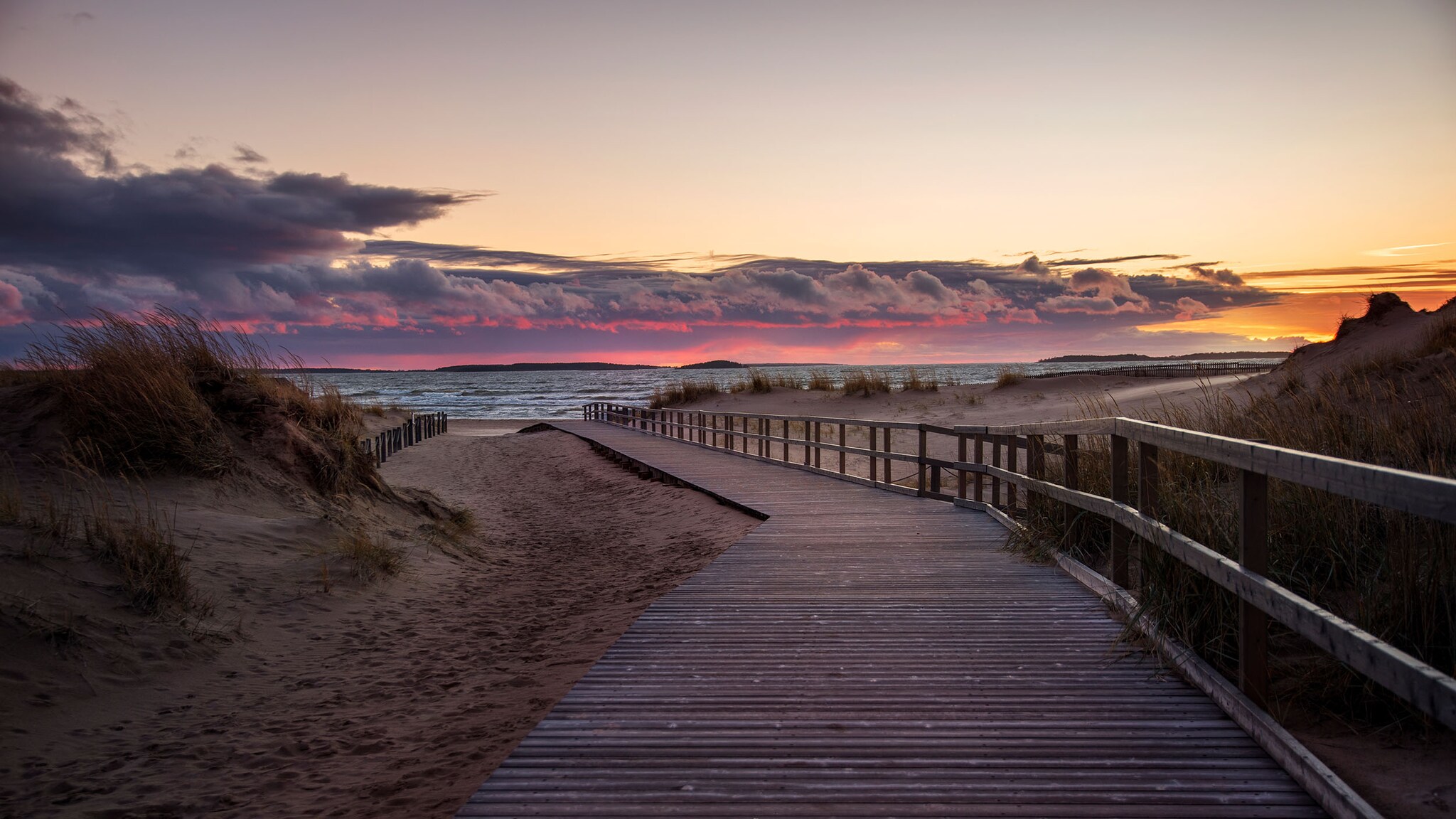 Ein breiter Holzsteg führt durch Dünen zu einem Sandstrand unter einem bunt gefärbten Himmel bei Sonnenuntergang.