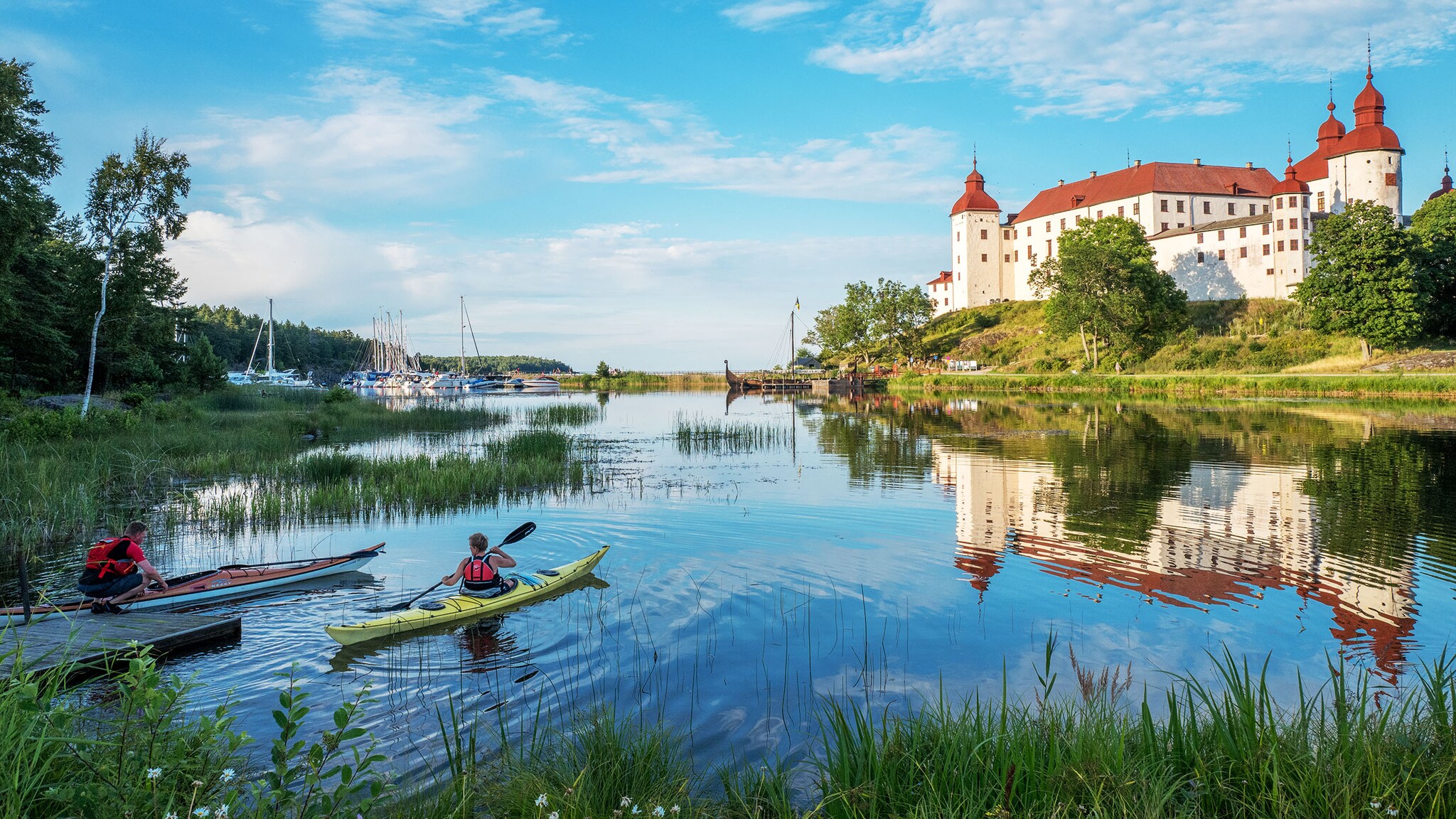 Zwei Personen befinden sich mit Kanus in einem See vor einem Schloss am grün bewachsenen Ufer bei Sonnenschein.