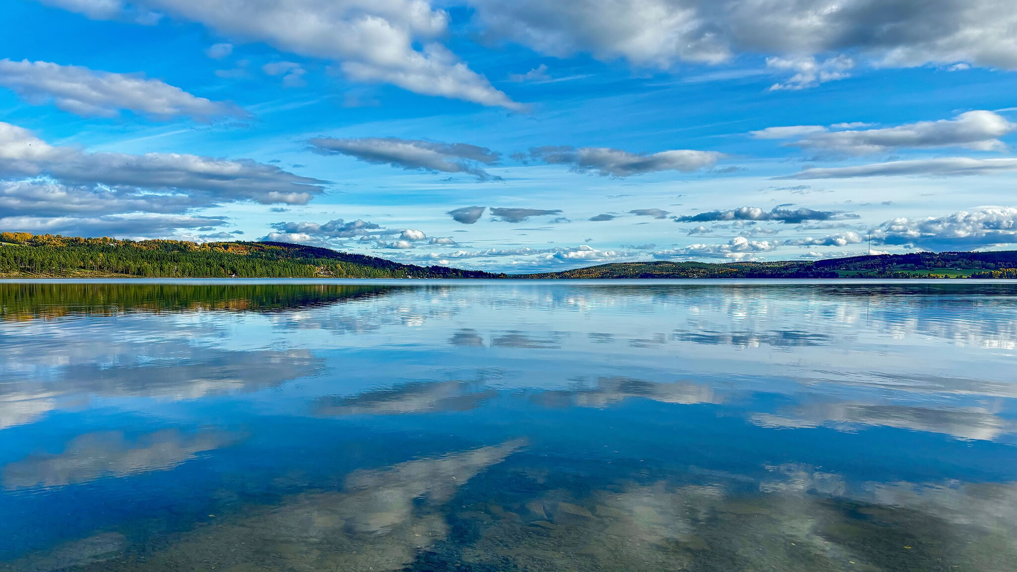 Weitläufiges Seepanorama mit Wolken am blauen Himmel, die sich auf der flachen Wasseroberfläche spiegeln.