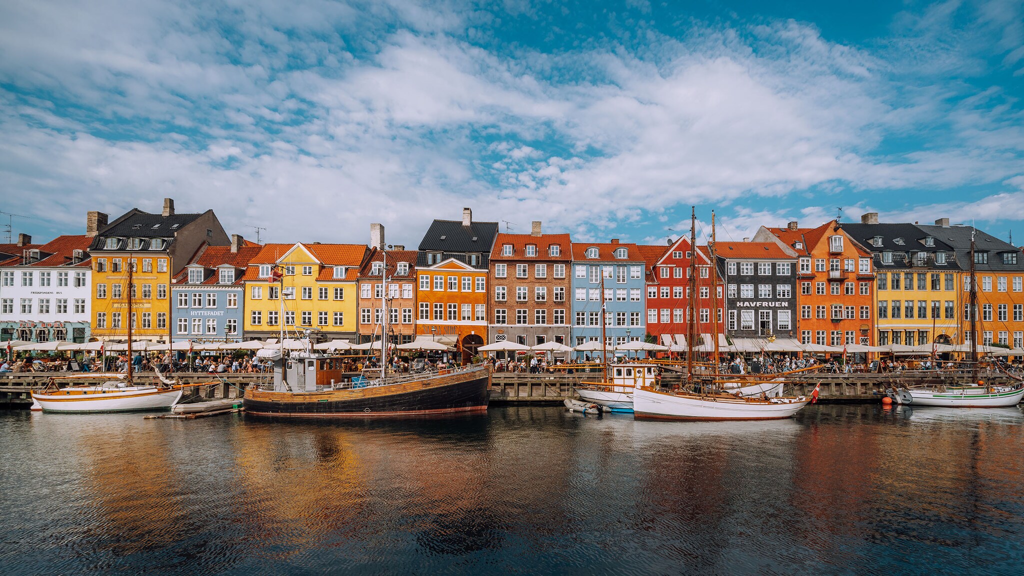Bunte Häuser mit Cafés an einer Promenade am Wasser mit historischen Holzschiffen.