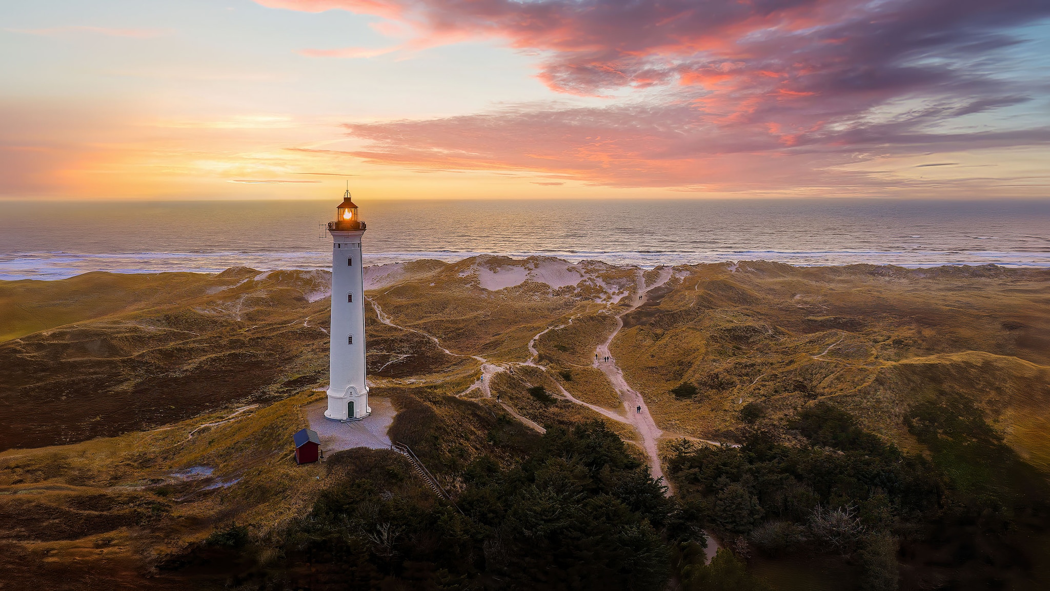 Weitläufige Dünenlandschaft mit Leuchtturm am Meer bei Sonnenuntergang.