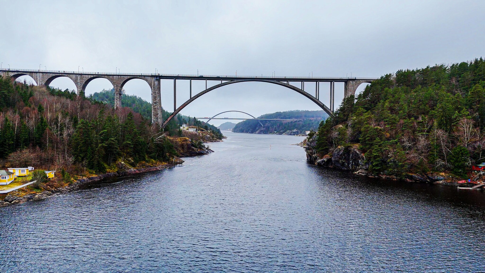 Zwei Brücken mit Rundbögen erstrecken sich über zwei bewaldete Ufer an einem Fjord unter grauem Himmel.