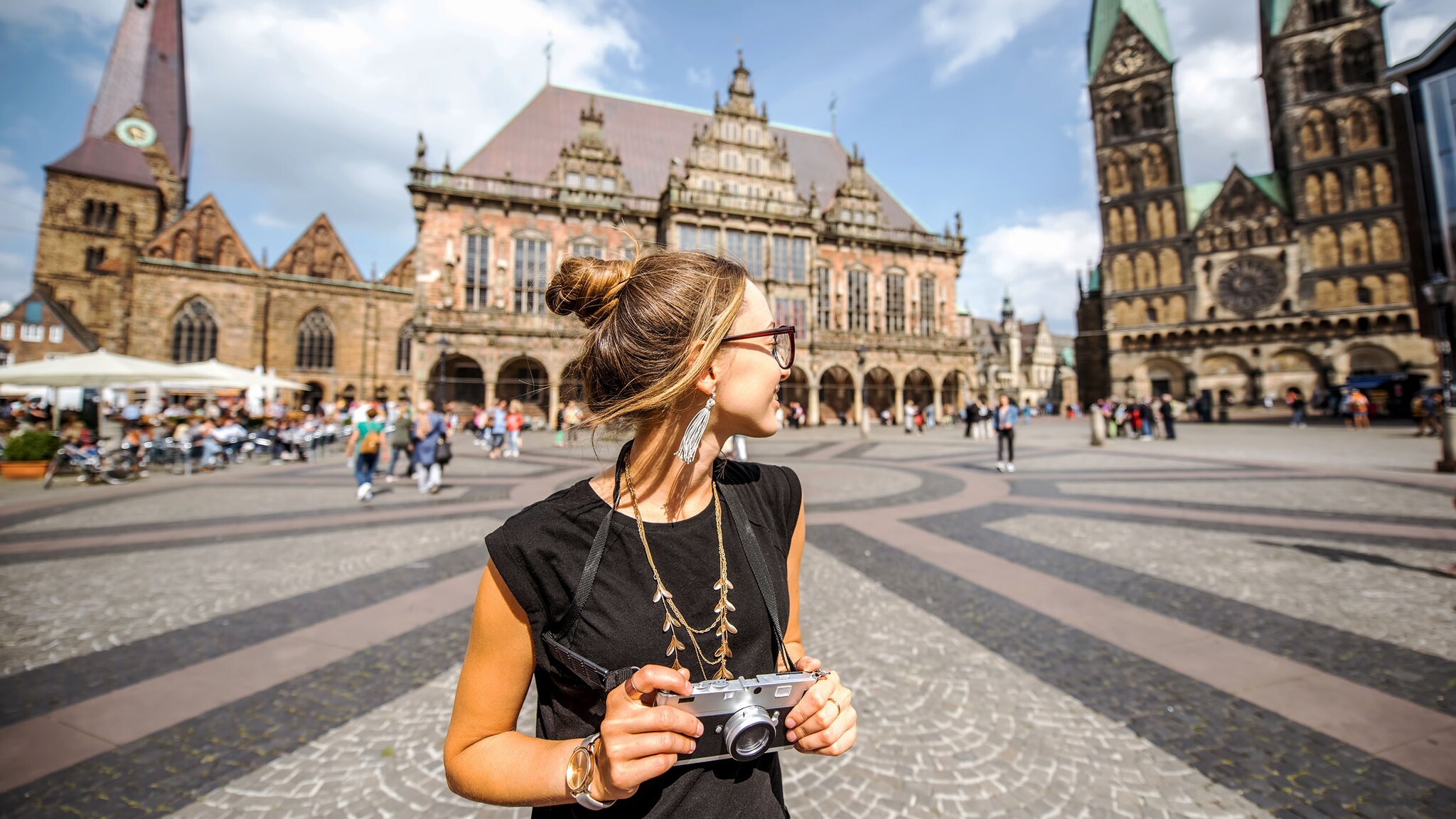 Eine Frau mit Sonnenbrille steht mit einer Kamera in den Händen auf dem historischen Bremer Marktplatz vor Rathaus und Kirche und blickt zur Seite bei Sonnenschein.