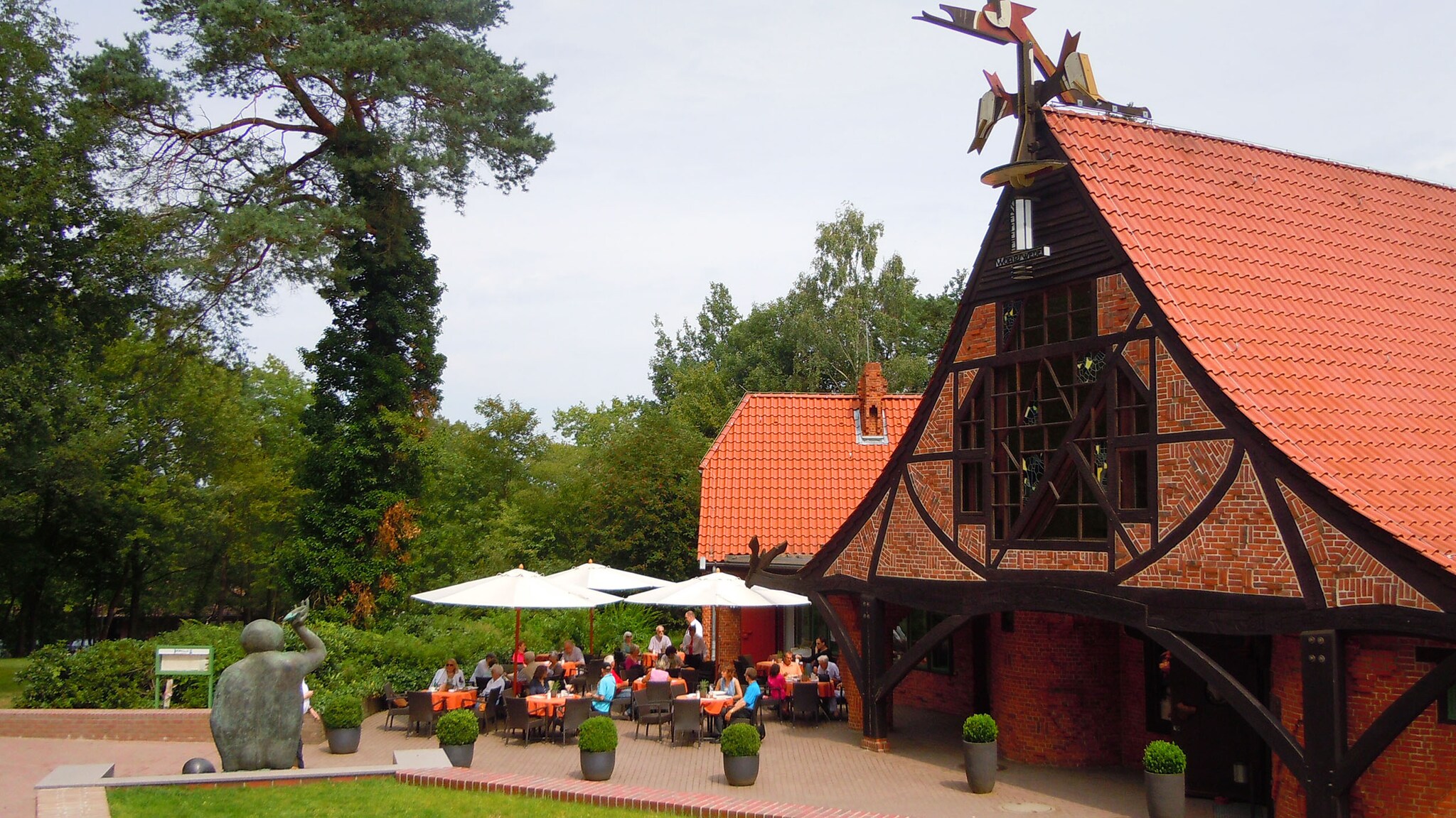 Personen sitzen auf der Terrasse eines Cafés an einem historischen Backsteinhaus mit tiefem, geschwungenen Dach in einer Parkanlage mit Skulpturen.