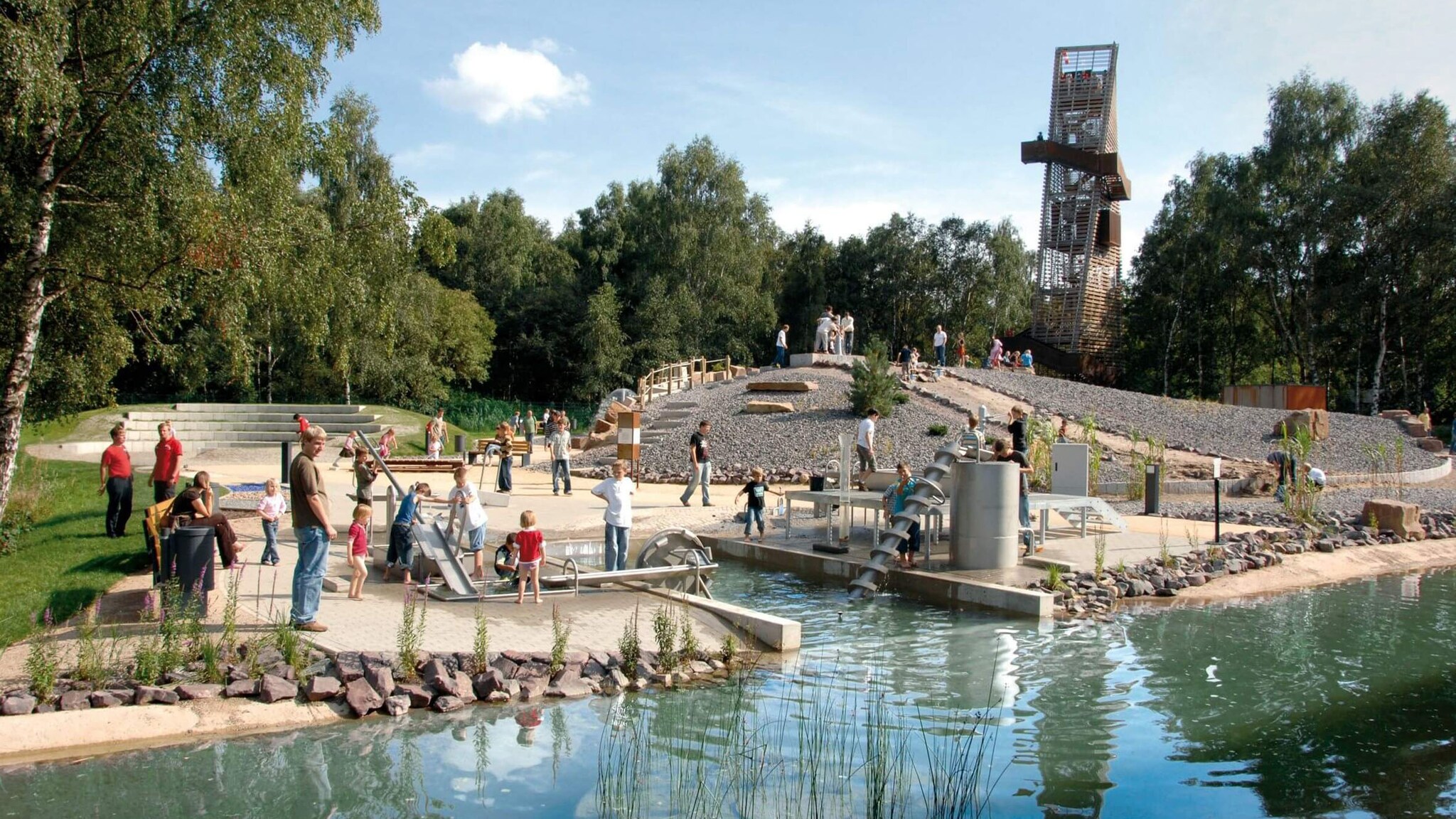 Erwachsene und Kinder spielen an Stationen auf einem Parkour auf einem Außengelände mit Wasserbecken bei Sonnenschein.