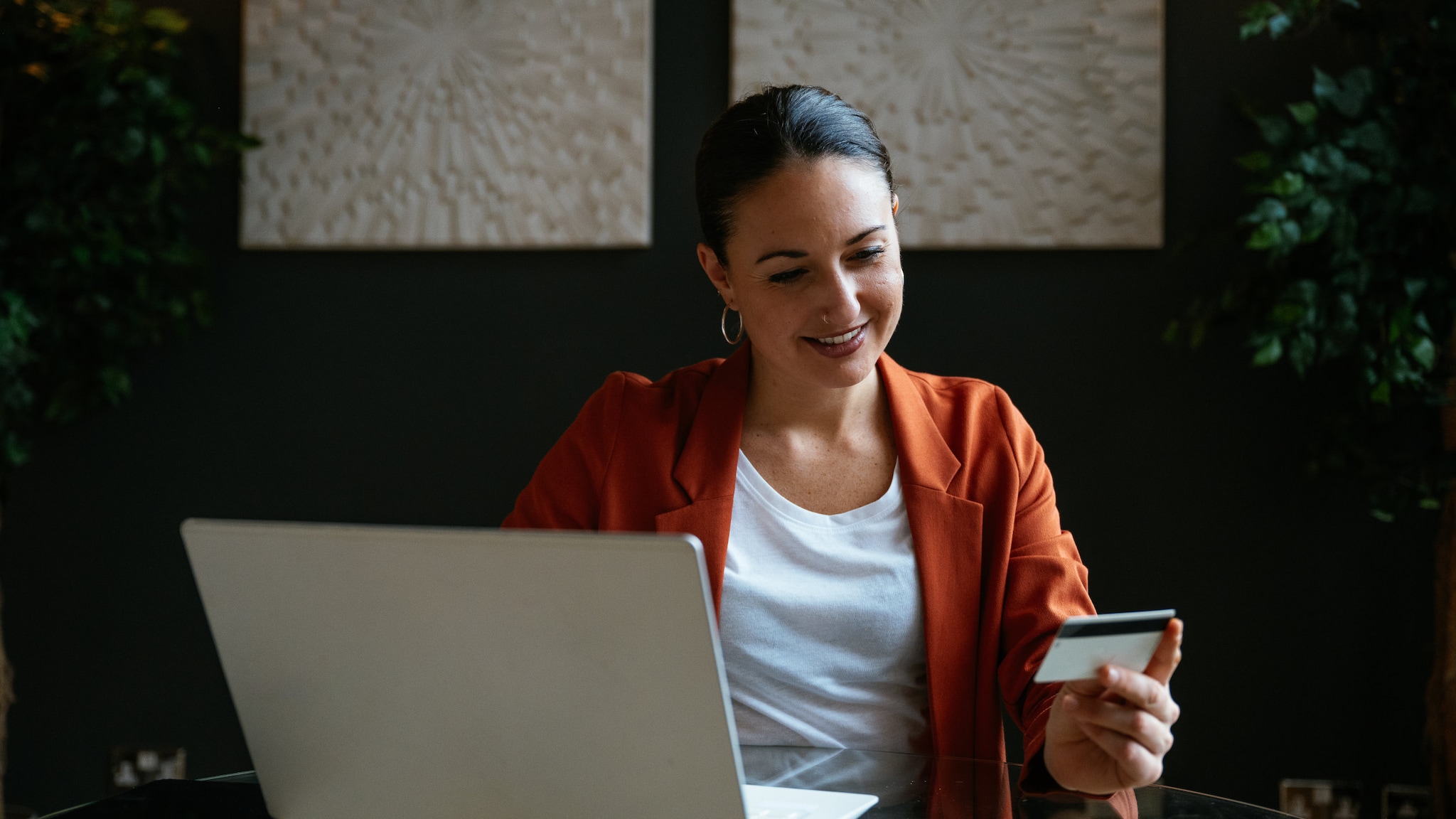 Eine lächelnde Frau sitzt vor einem aufgeklappten Laptop und schaut auf eine Bankkarte in ihrer Hand.
