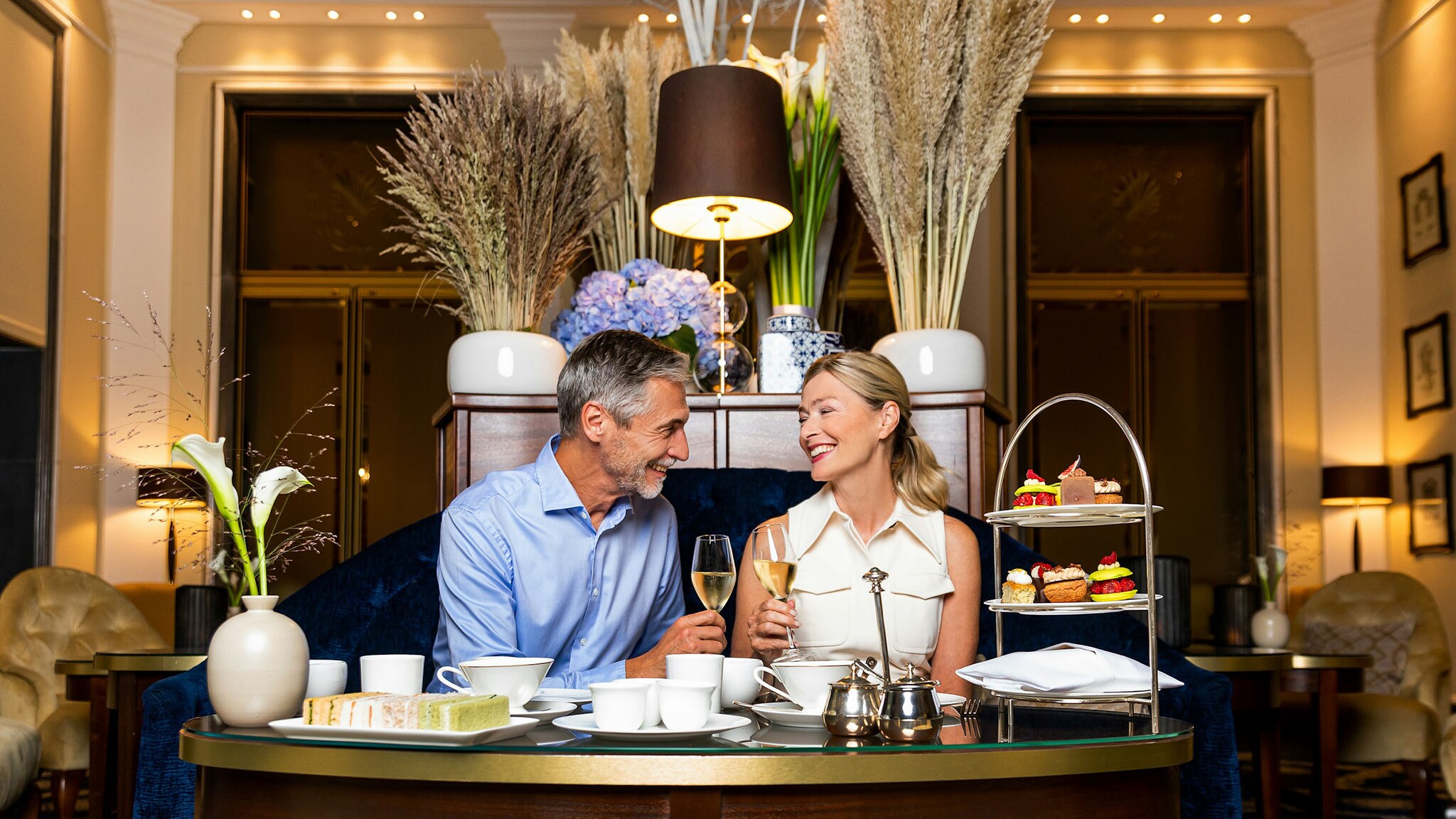 Ein Mann und eine Frau sitzen mit Champagnergläsern in den Händen an einem Tisch beim Afternoon-Tea in einem eleganten Salon.