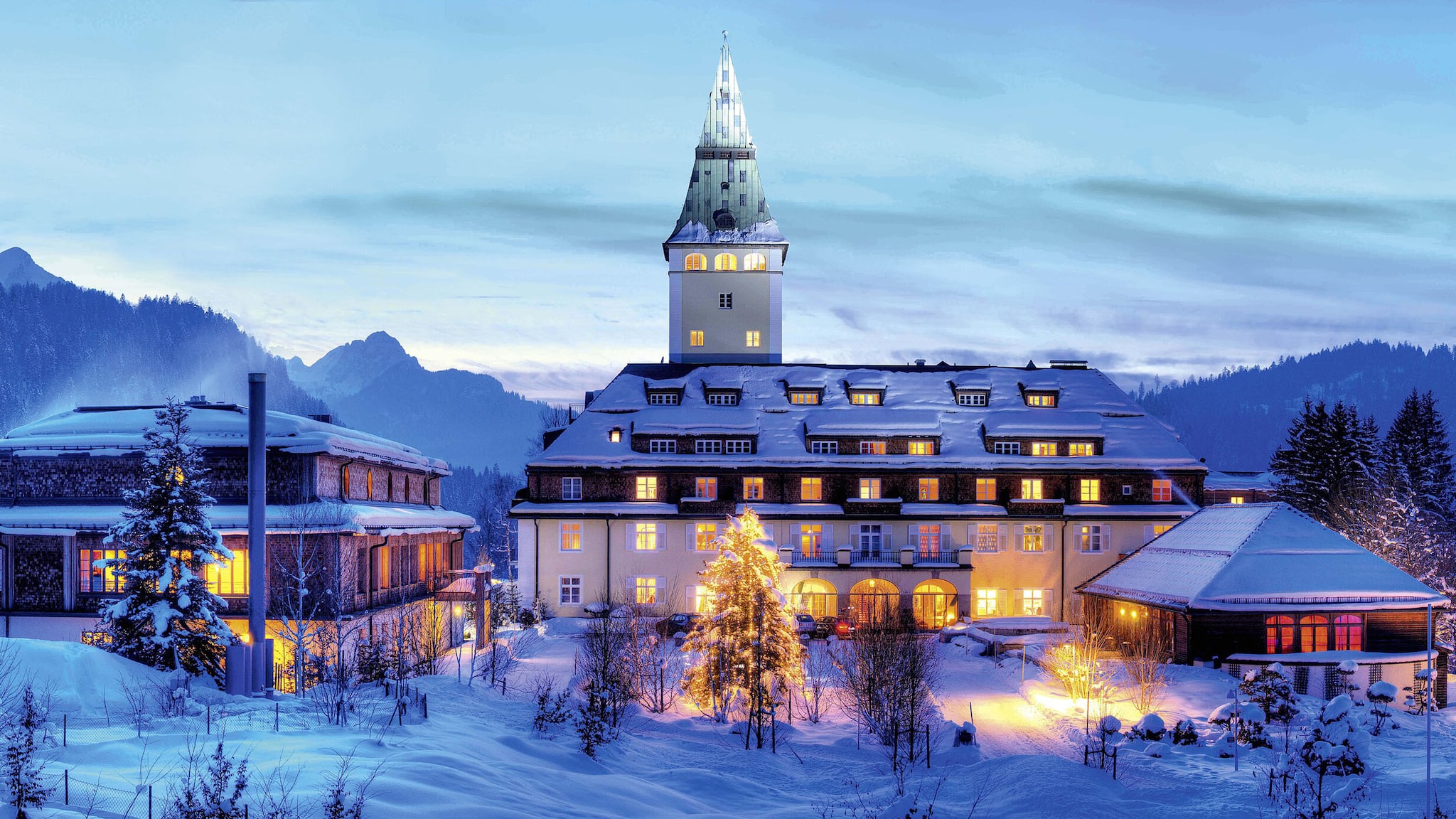 Ein beleuchteter, schlossartiger Hotelkomplex in einer Schneelandschaft vor Bergpanorama bei Abenddämmerung.