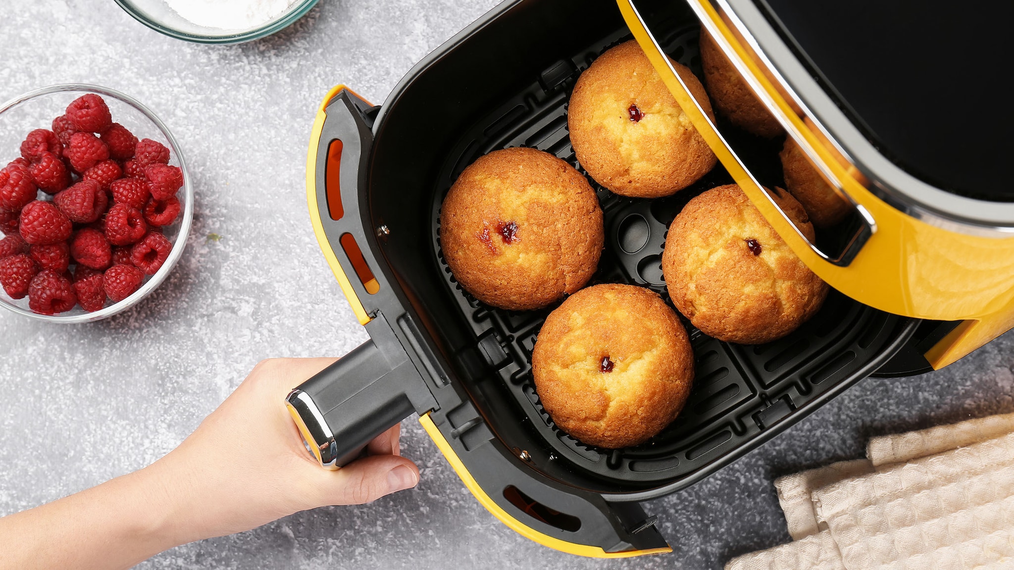 Aufsicht einer Hand am Behälter eines Airfryers, gefüllt mit vier runden Kuchen, auf einem Küchentisch mit Zutaten.