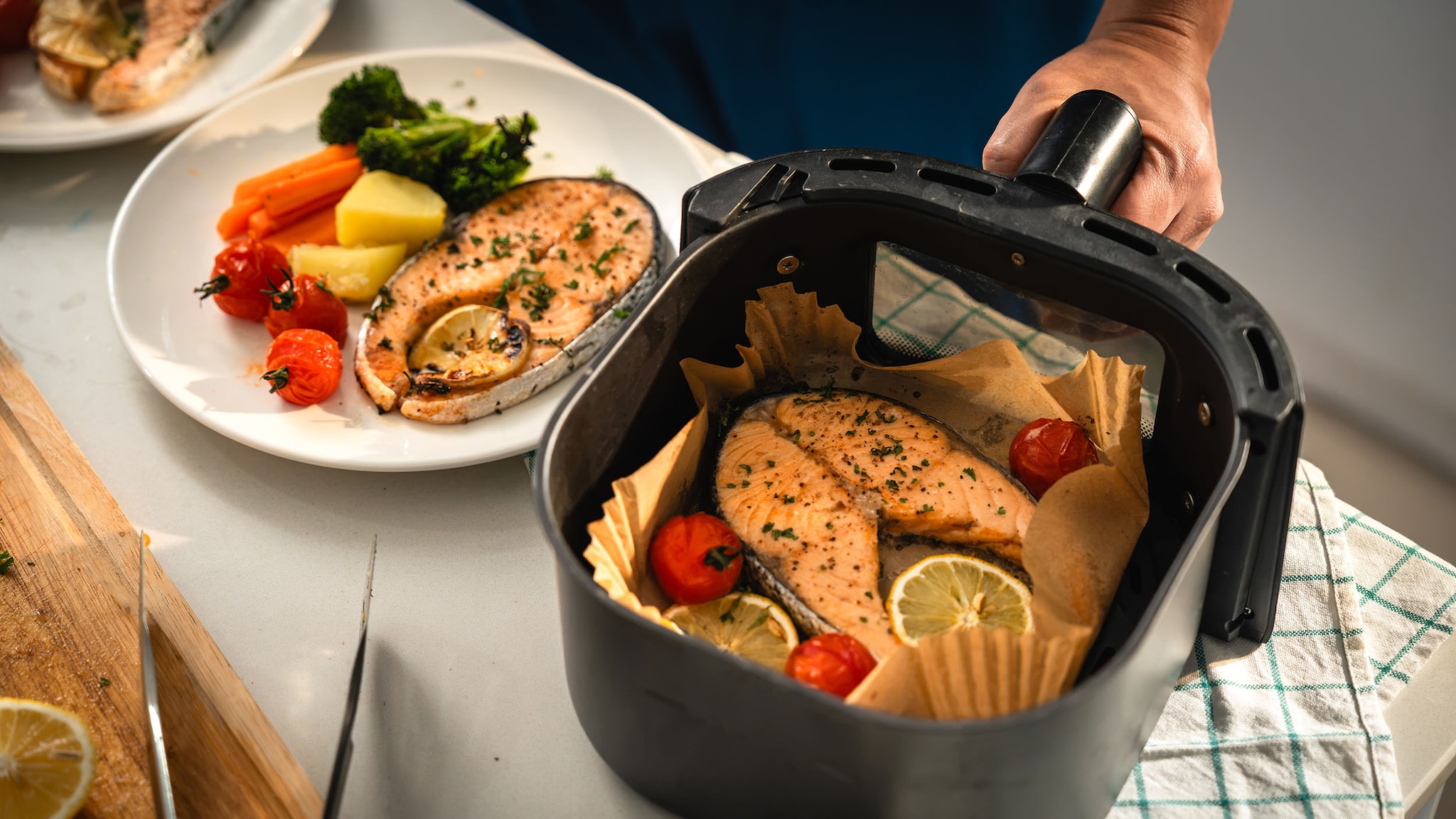 Ein Teller und ein Behälter eines Airfryers mit Lachsfilets und Gemüse stehen auf einer Küchentheke.