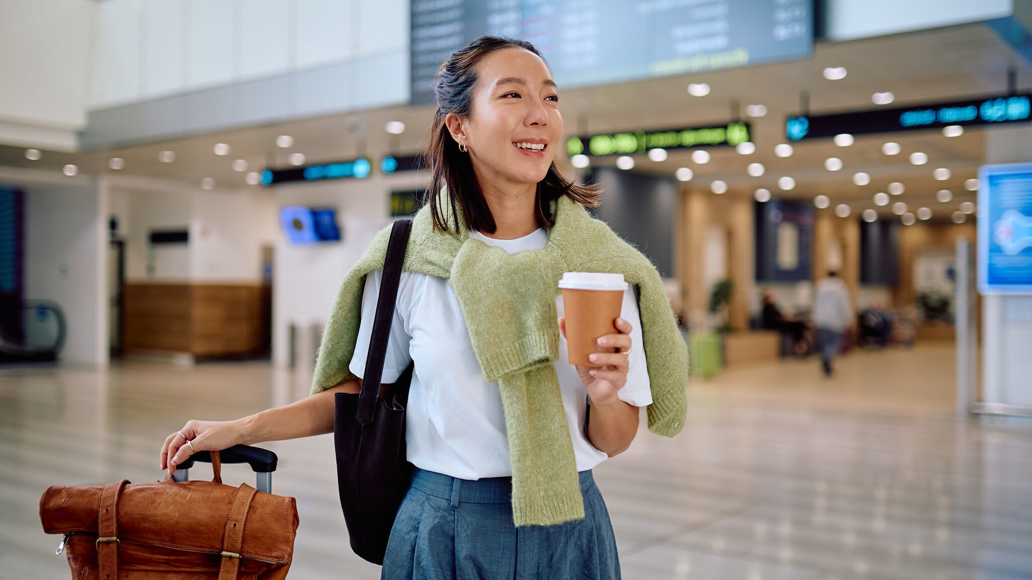 Eine lächelnde Frau steht mit Reisegepäck und einem Kaffeebecher in der Hand in einem Flughafenterminal.