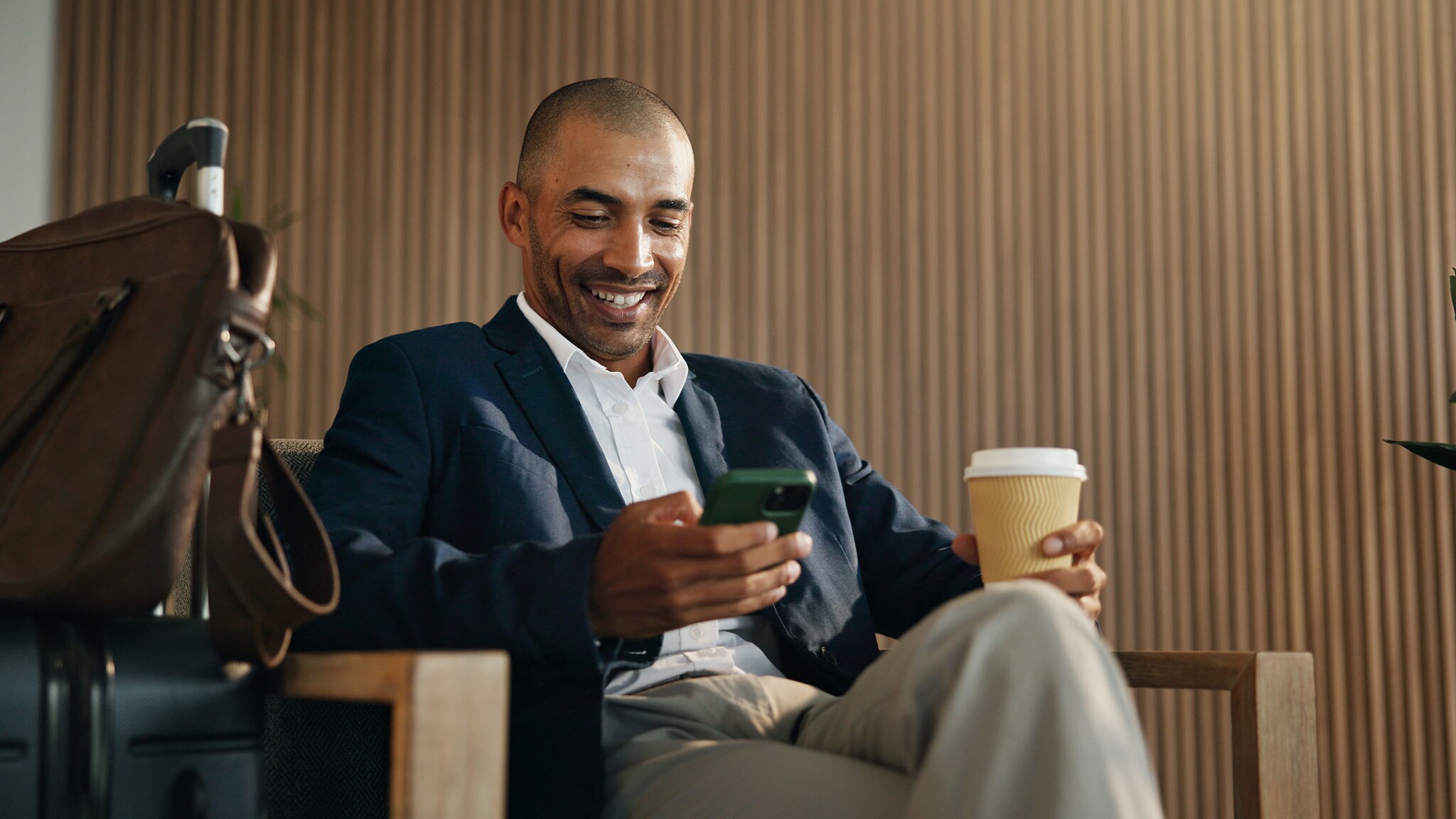 Ein elegant gekleideter Mann sitzt in einem Sessel und schaut lächelnd auf ein Smartphone in seiner Hand, in der anderen hält er einen Kaffeebecher, neben ihm steht Reisegepäck.