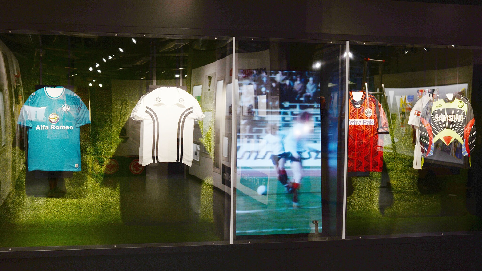 Verschieden Fußballtrikots hängen in einer Vitrine neben einem Display, das ein Fußballsiel zeigt.