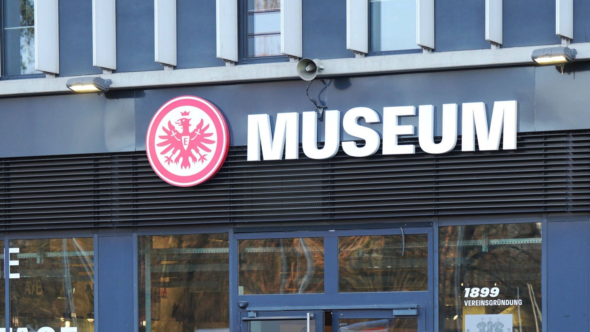 Moderne Außenfassade eines Museums mit rundem, rotem Adlerwappen über dem Eingang.