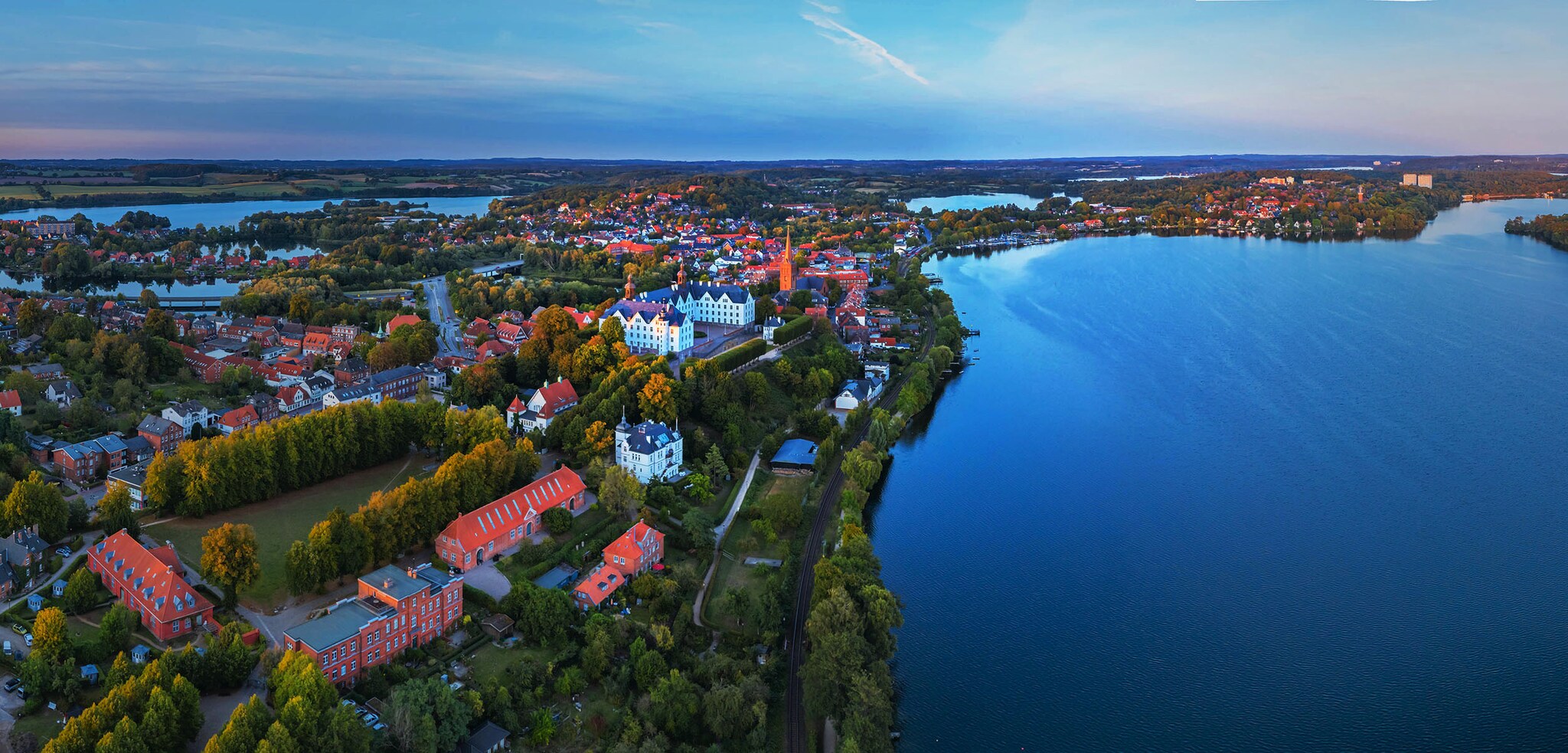Luftaufnahme einer Seenlandschaft mit einer Stadt mit Schloss und Waldgebieten am Wasser bei Abenddämmerung. 