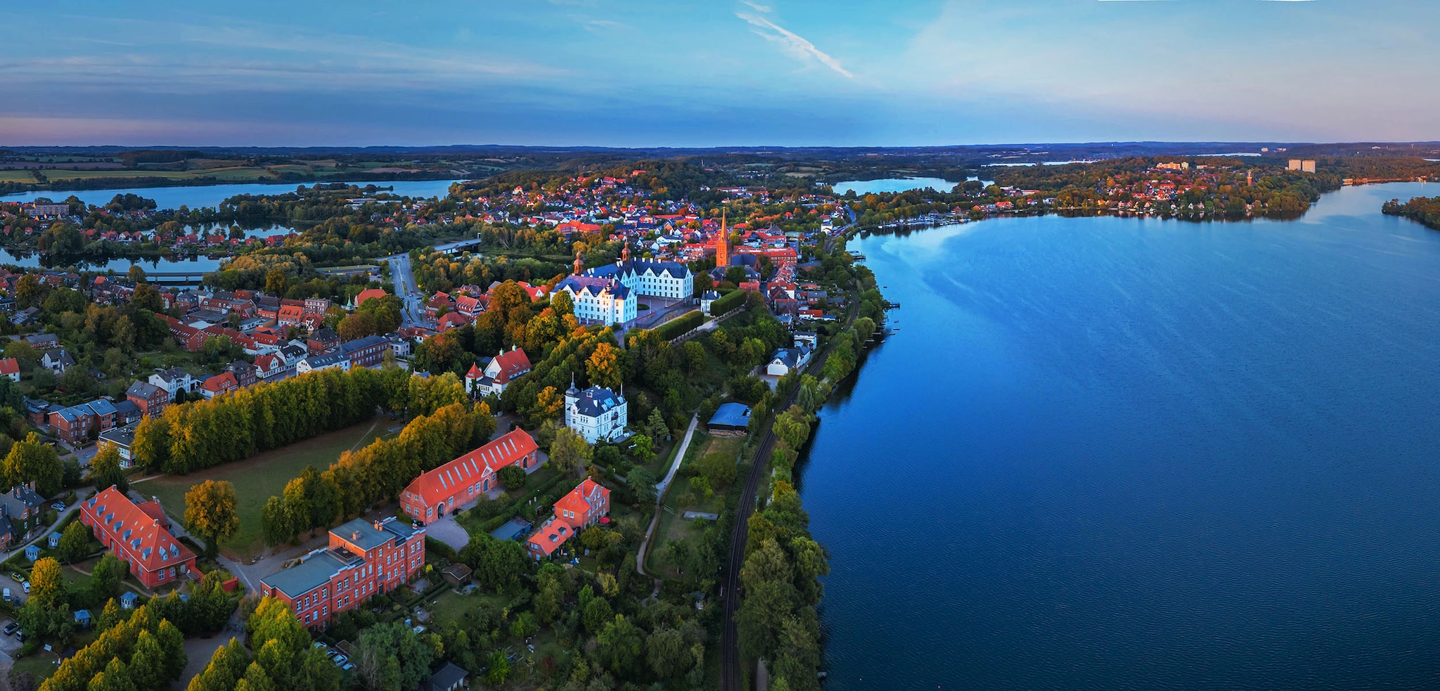 amex-magazin-schleswig-holstein-lieblingsorte-ploen Luftaufnahme einer Seenlandschaft mit einer Stadt mit Schloss und Waldgebieten am Wasser bei Abenddämmerung.