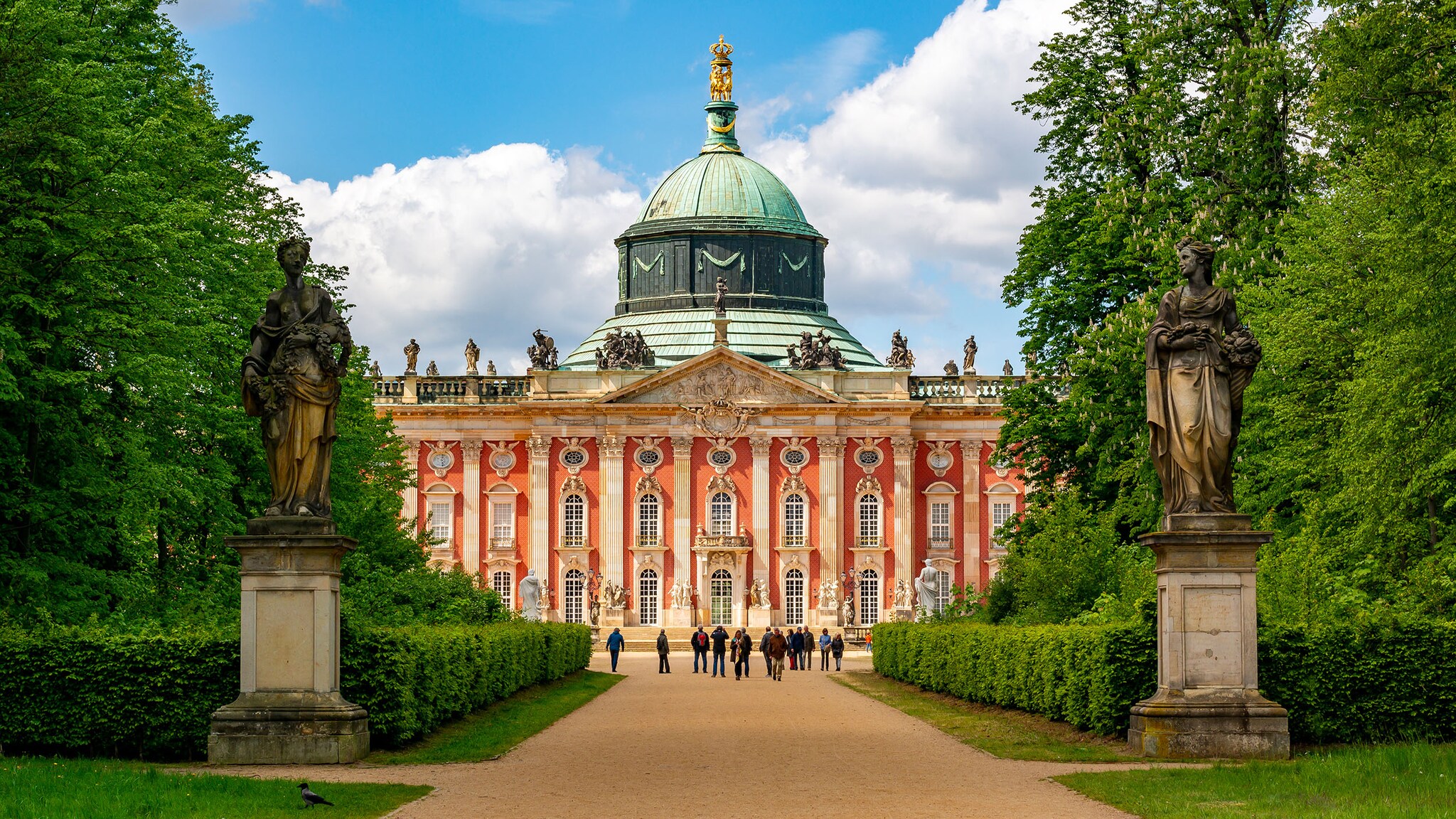 Personen vor einer barocken Schlossanlage im Park Sanssouci, im Vordergrund flankieren Statuen den Weg. 
