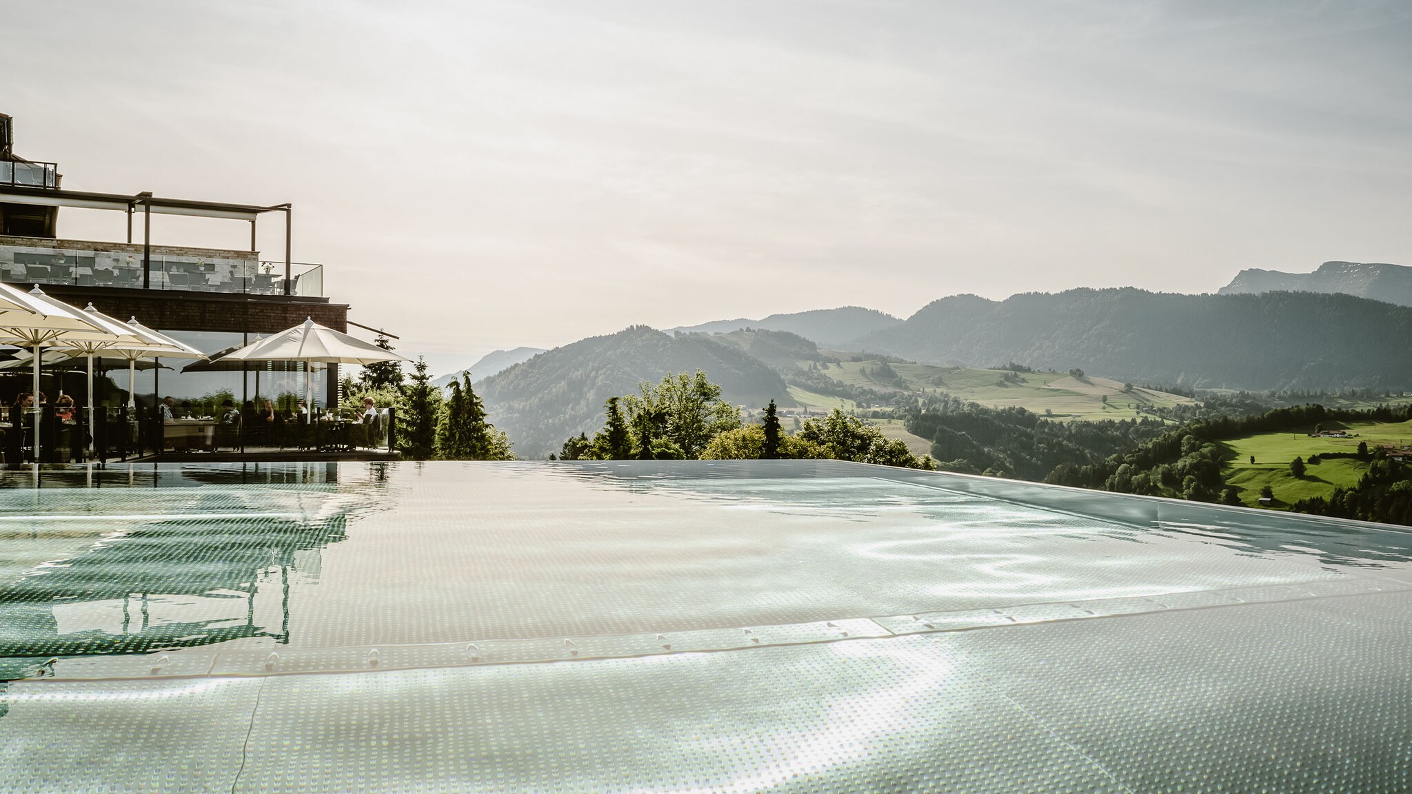 Infinity-Pool an einem modernen Hotelkomplex, umgeben von grüner Natur vor Bergpanorama.