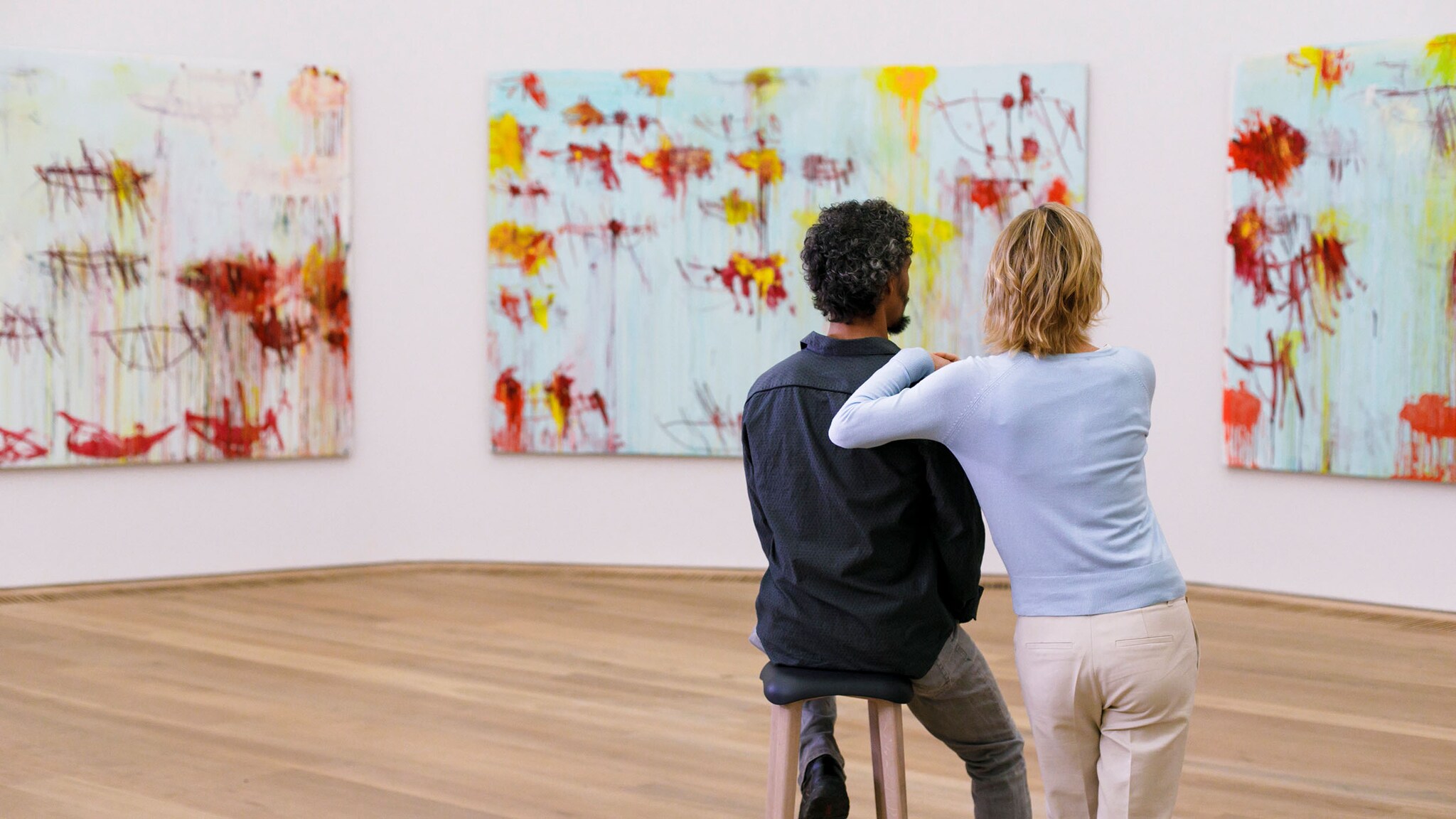 Rückansicht von zwei Personen in einem hellen Ausstellungsraum vor drei Gemälden mit abstrakter Kunst.