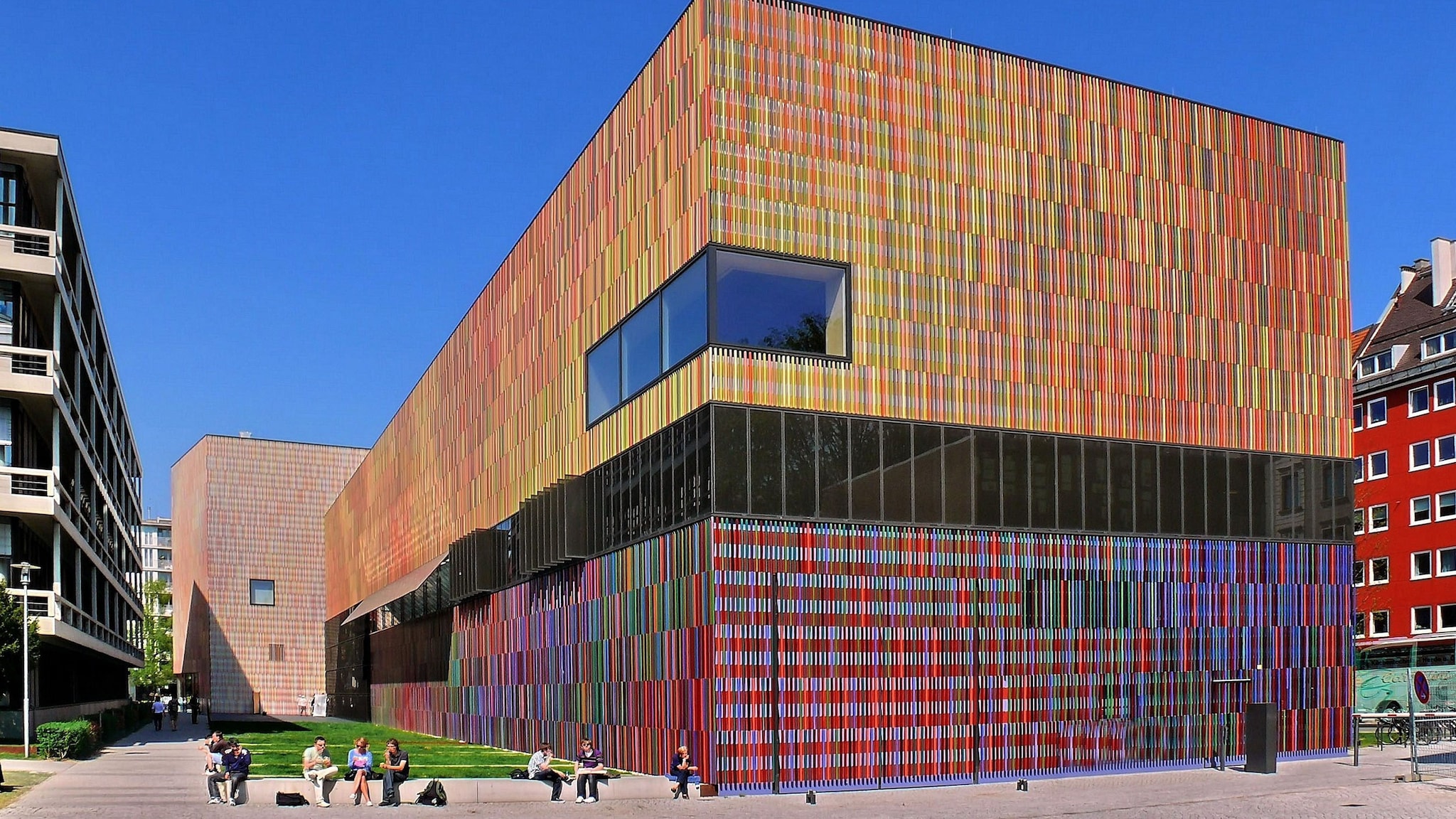 Personen im Freien an einem modernen, kubusförmigen Museumsgebäude mit einer Fassade aus bunten Keramikstäbchen unter blauem Himmel.