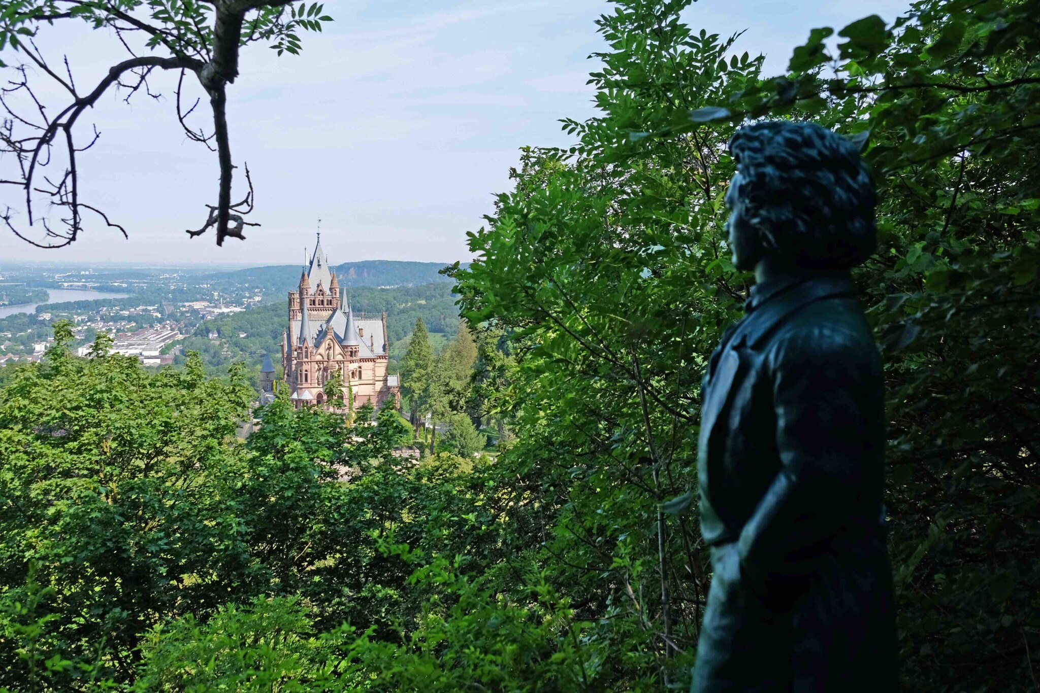 Ein altes Schloss auf einer Anhöhe, im Tal der Rhein, im Vordergrund eine Beethoven Statue und zwischendrin viele grüne Bäume und Sträucher