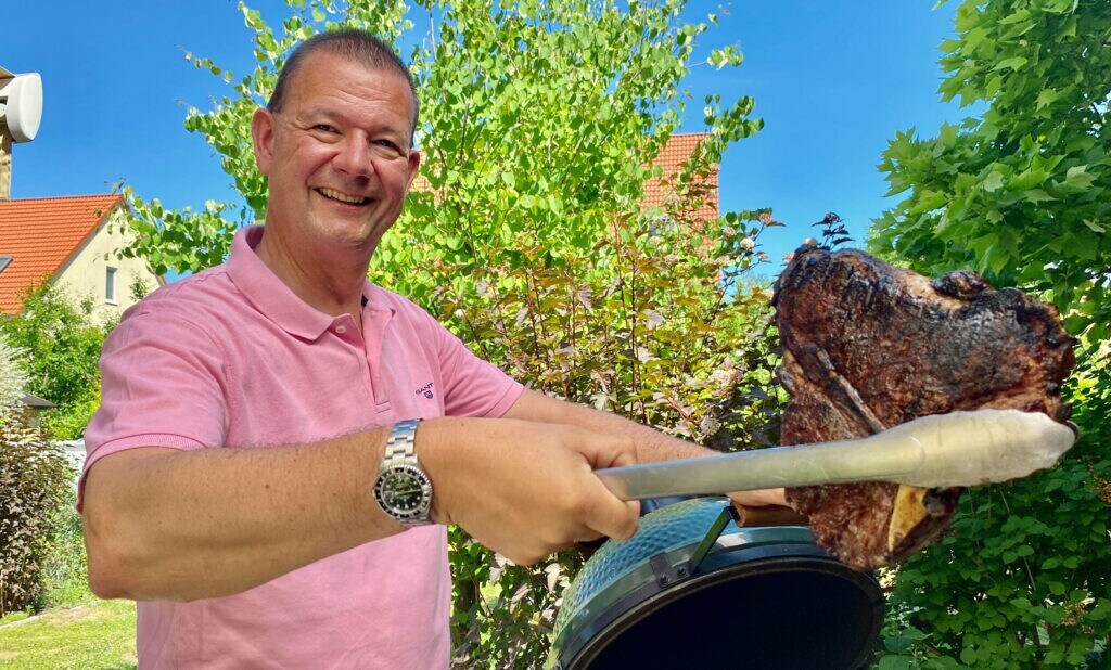 Ein Mann steht an einem Kugelgrill und hält ein fertiges Steak in der Grillzange