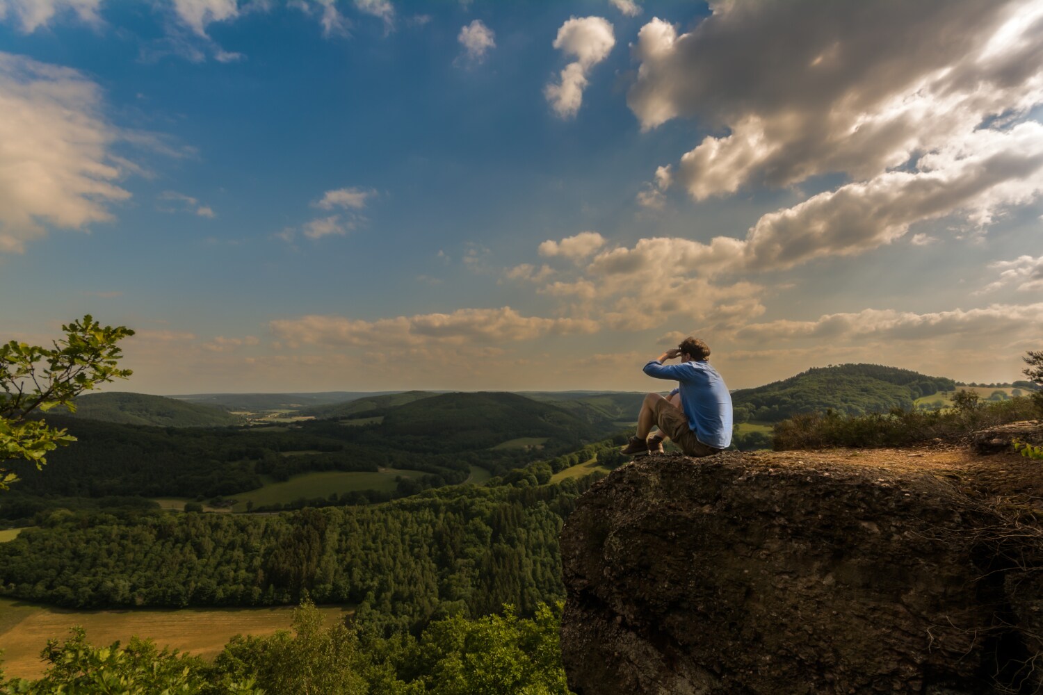 Ein Mann sitzt auf einem Felsen und blickt über die grüne Landschaft der Eifel