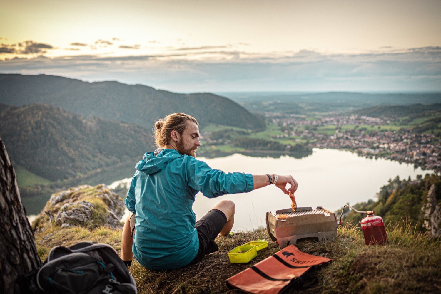 Ein Mann sitzt auf einem Berg mit Blick auf einen See während er ein Stück Fleisch auf den Grill legt