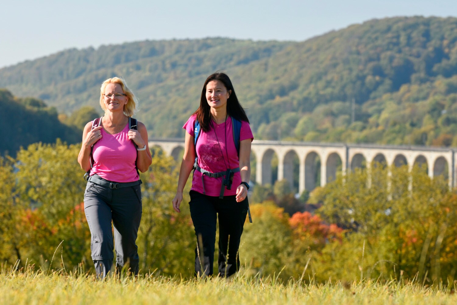 Zwei Frauen wandern durch ein hügelige, waldige Landschaft, im Hintergrund der Viadukt